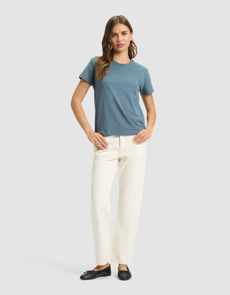 A woman with long, wavy hair stands facing forward against a plain light gray background, wearing the Cozy Earth Women's Easy Tee in blue, off-white pants, and black slip-on shoes, with her hands resting on her hips.