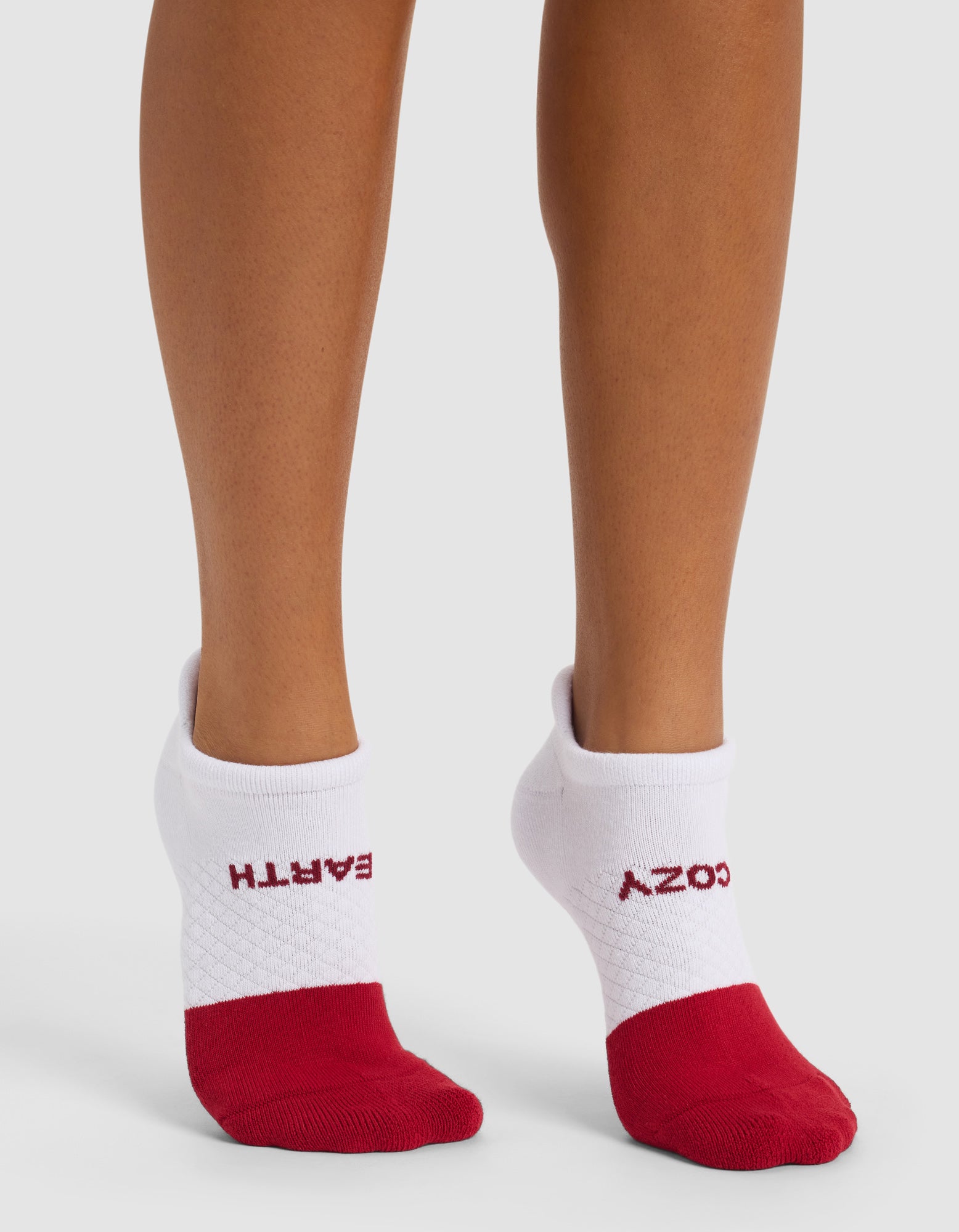 A person wears Cozy Earth Essential Ankle Socks—white with red toes and heels. "EARTH" is printed on the left cuff, while "COZY" appears upside down on the right sock.|Color:Crimson
