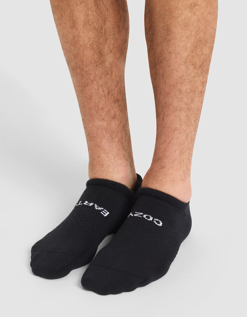 A person wearing Cozy Earth Essential Ankle Socks in black with white text stands on a light gray floor, showing their lower legs and feet partially covered by the socks.