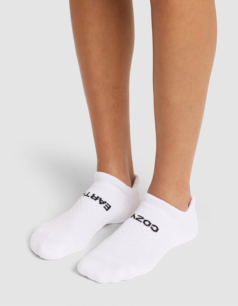 A person wears Cozy Earth Essential Ankle Socks with black text atop, standing on a light gray surface; only their lower legs and feet are shown.