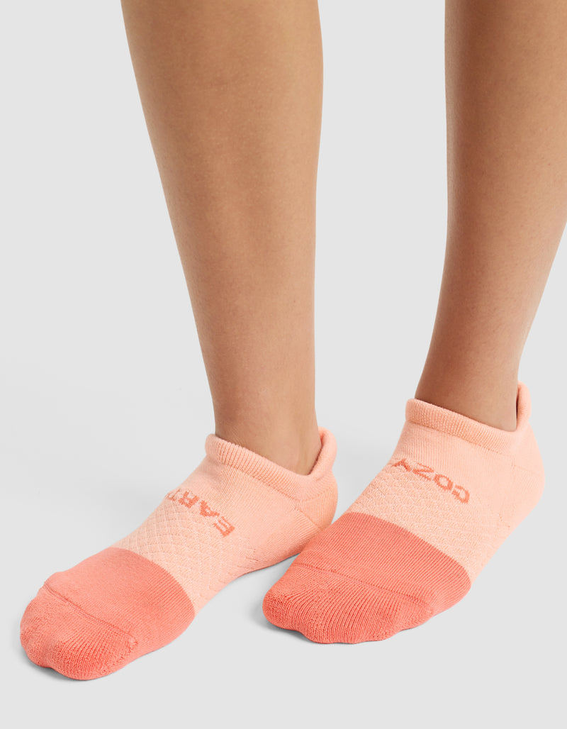 A person is wearing Cozy Earth's Essential Ankle Sock in light pink with a darker pink toe, standing on a light surface. Only their lower legs and feet are visible.
