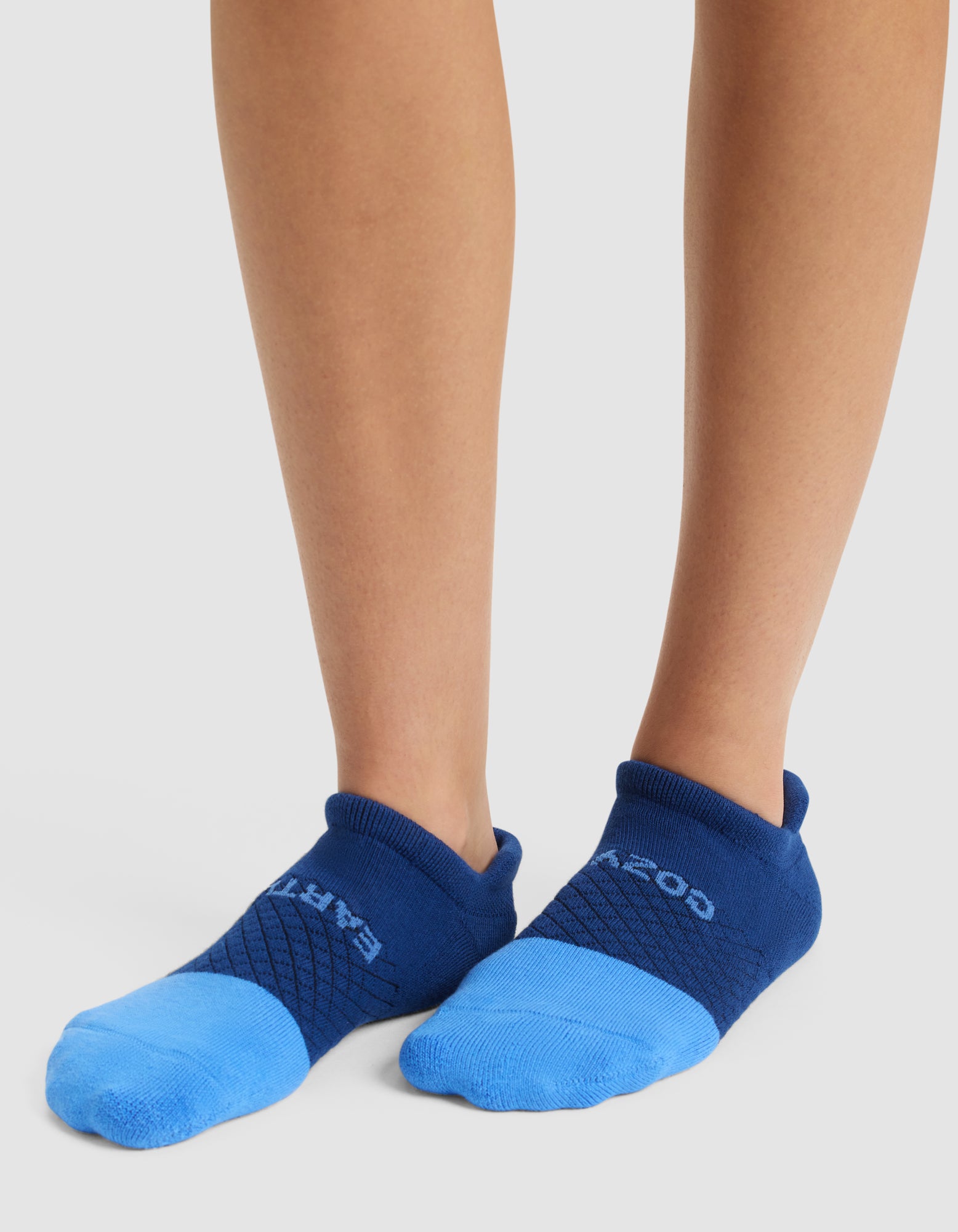 A person stands on a white surface wearing Cozy Earth Essential Ankle Socks in blue with darker blue cuffs and accents; "EARTH" appears on the left sock and "GOOD" on the right. |Color:Harbor