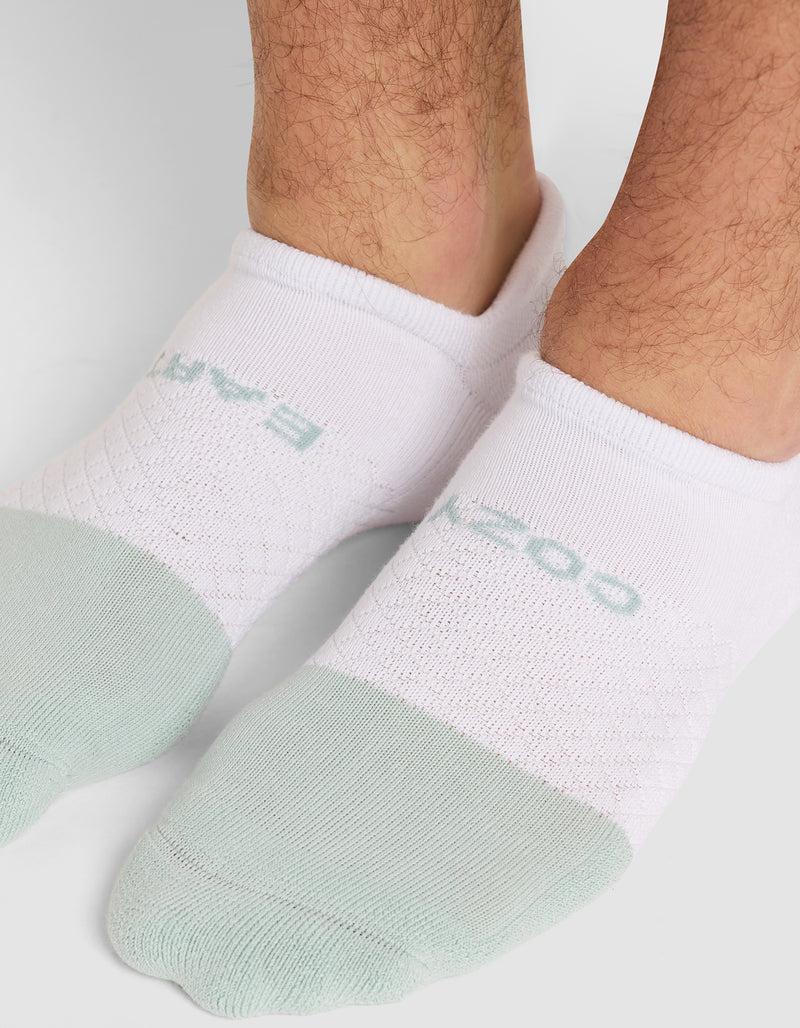 Close-up of a person's lower legs and feet wearing Cozy Earth Essential Ankle Socks with light green toes and light blue text on top, standing on a white surface.