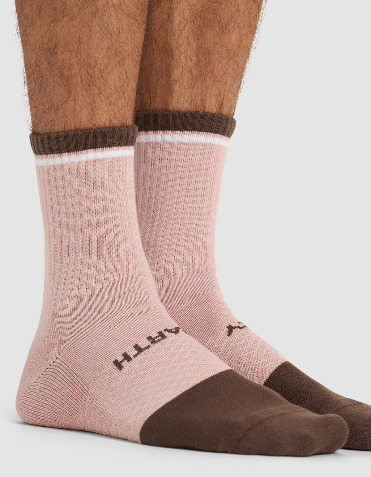 Two feet wearing Cozy Earth Men’s Essential Calf Socks in a pink ribbed design with brown toes, heels, and cuffs. The socks feature a thin white stripe near the top and branded text across the toes. The background is plain and light-colored.