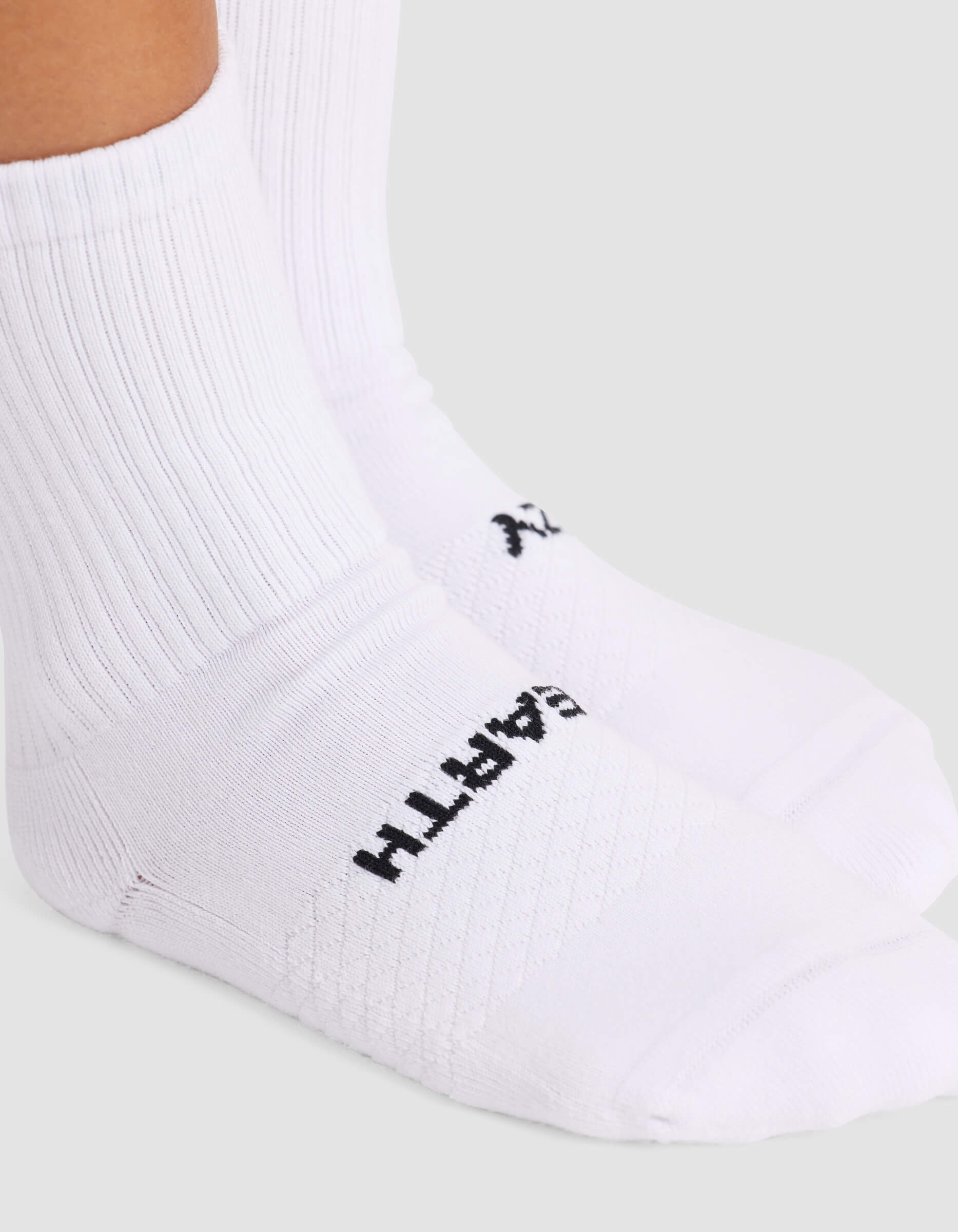 A close-up shows a person wearing Cozy Earth Women’s Essential Calf Socks in white with ribbing and a logo near the toes. The background is plain and light-colored.