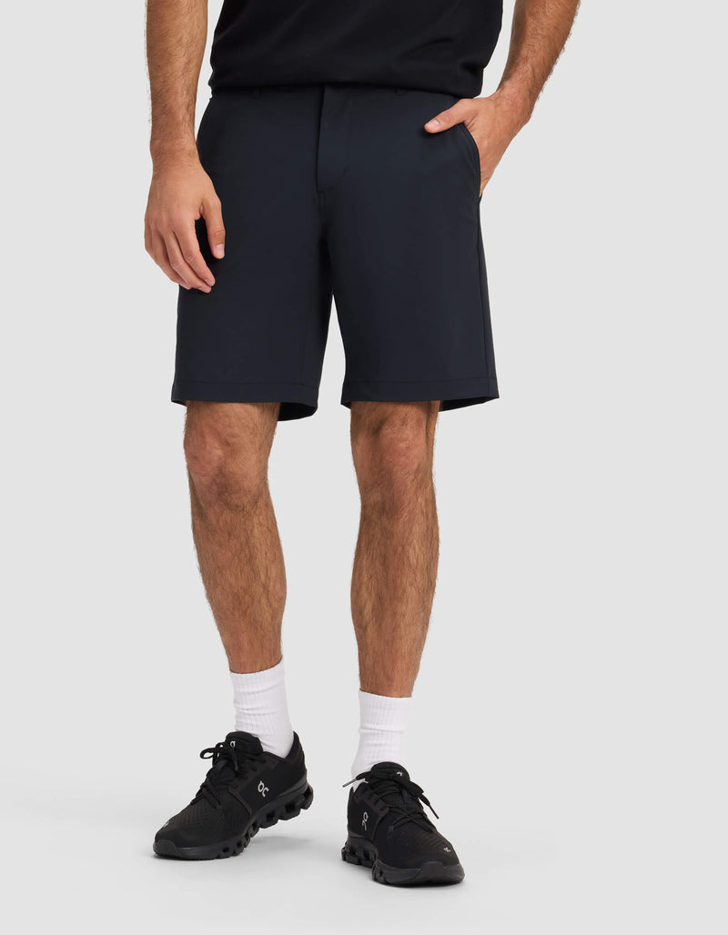 A man wearing Cozy Earth's Men's Everywhere Short in black, a black shirt, black sneakers, and white crew socks stands with one hand in his pocket against a plain light gray background.