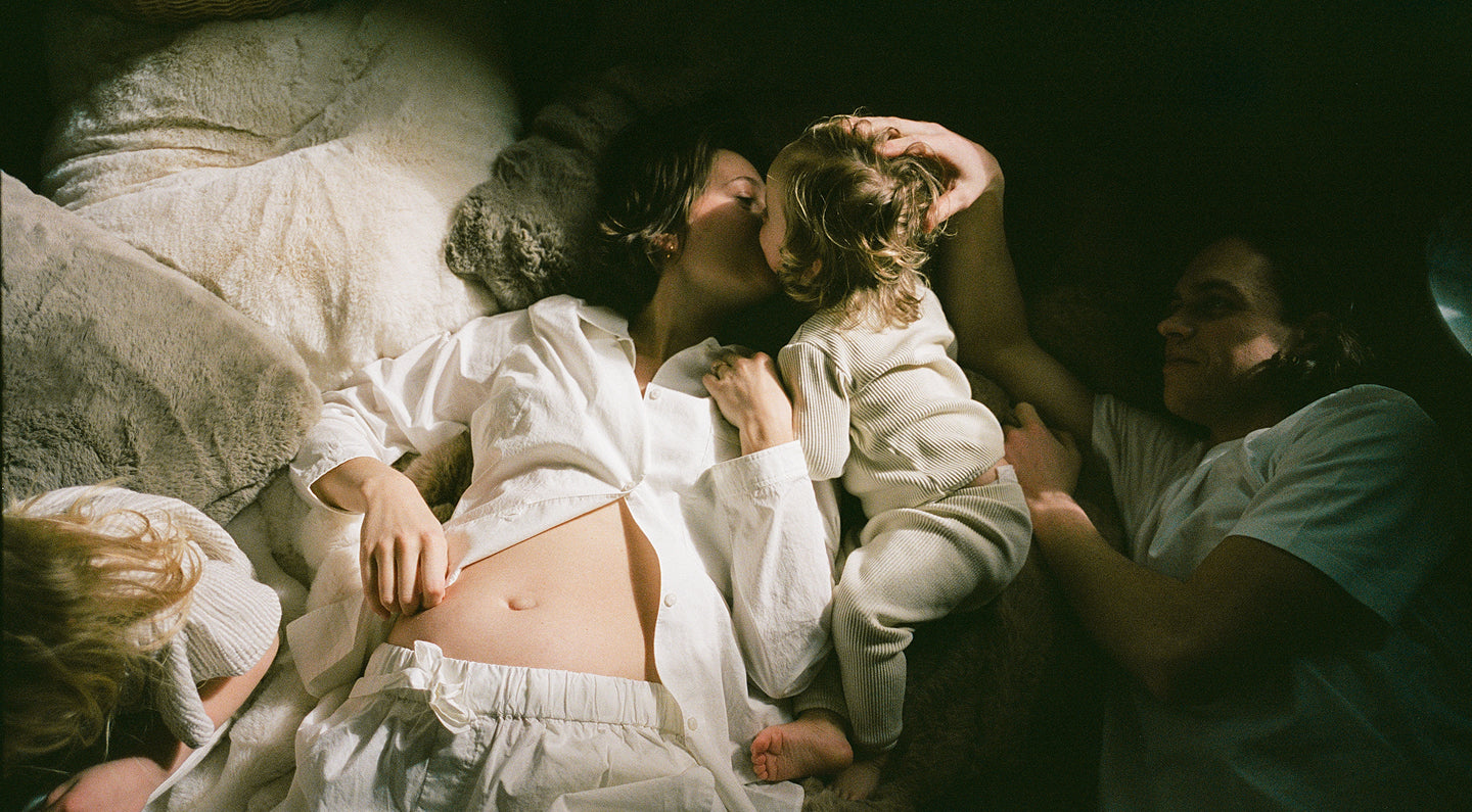 A warm, high-contrast lifestyle photograph of a family lying together on a bed. A woman in a white button-down shirt and matching sweatpants holds a young child wearing a cream-colored long-sleeve top and matching bottoms.
