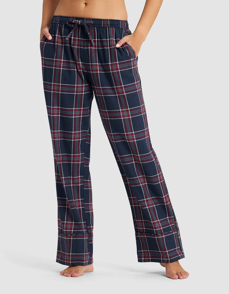 A person stands barefoot with hands in pockets, wearing Cozy Earth Women's Farmhouse Pajama Pant in a navy blue, red, and white plaid pattern. Their upper body is partially out of frame against a plain background.