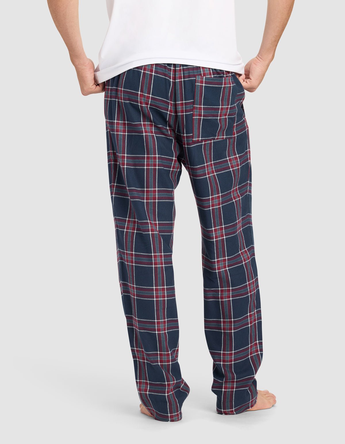 A person stands barefoot, facing away, wearing a white shirt and Cozy Earth Men’s Farmhouse Pajama Pant in navy blue with a red and white plaid pattern. The background is plain light gray.|Color:Eclipse Plaid