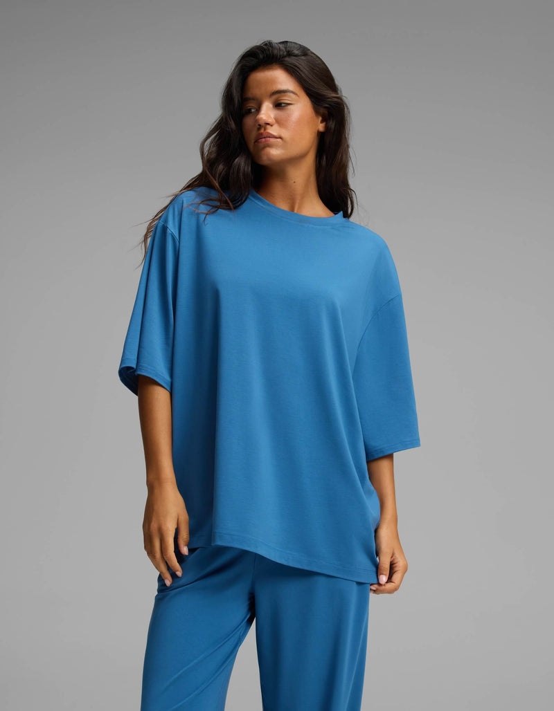 A woman with long brown hair wears Cozy Earth’s Women’s First Layer Boyfriend Sleep Tee in blue, paired with matching pants. She stands against a plain gray background, gazing slightly to the side with a neutral expression.