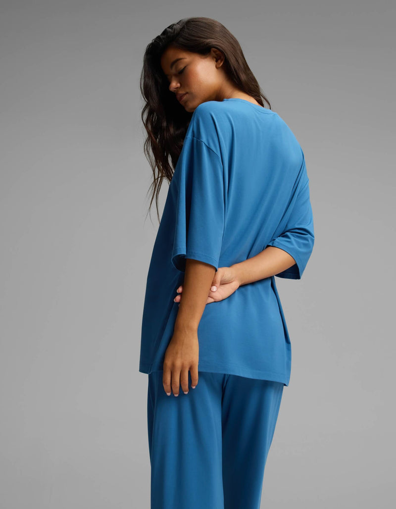 A person with long dark hair stands in profile against a plain gray background, wearing the Cozy Earth Women’s First Layer Boyfriend Sleep Tee in blue, paired with matching pants. One arm is bent behind their back.