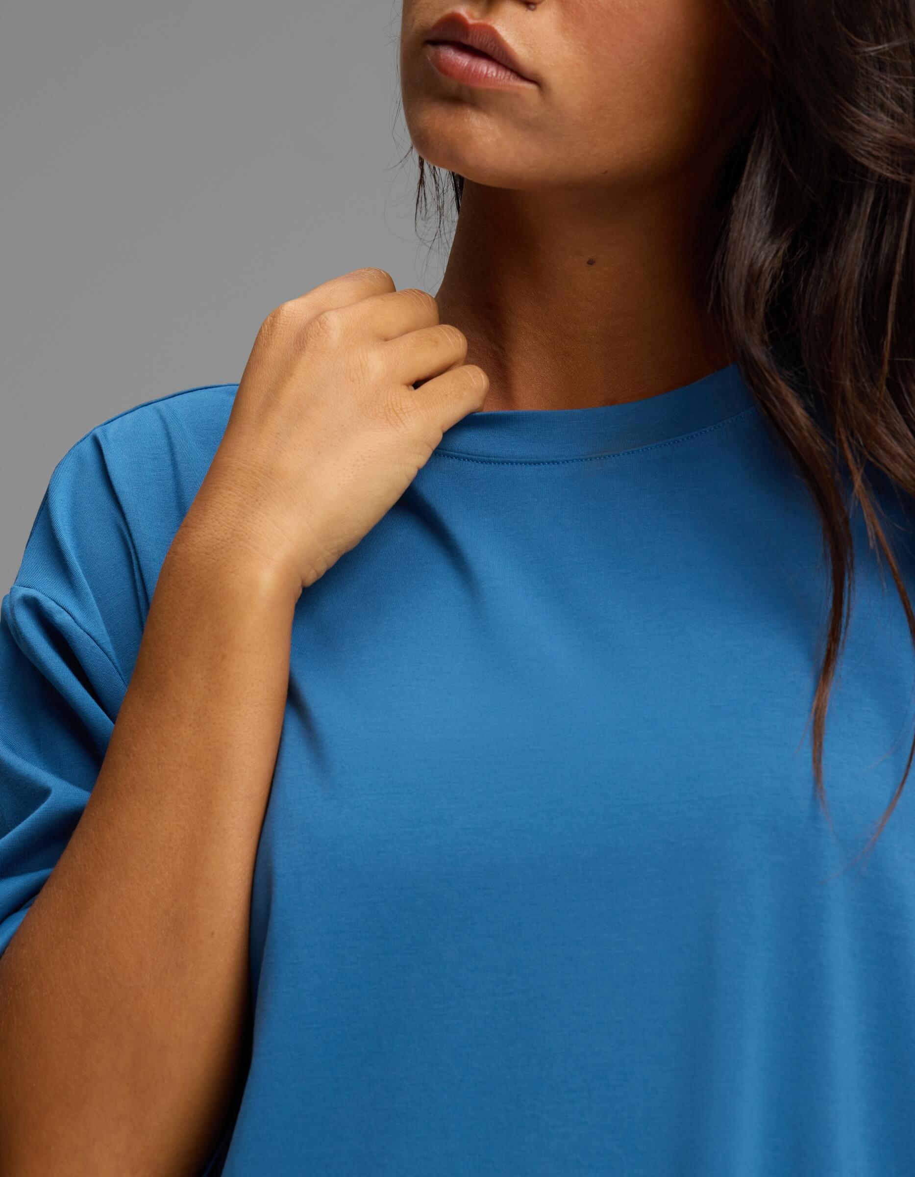 A person wearing the Cozy Earth Women’s First Layer Boyfriend Sleep Tee in blue touches the collar with one hand. The lower part of their face and dark hair are visible against a plain, neutral background.