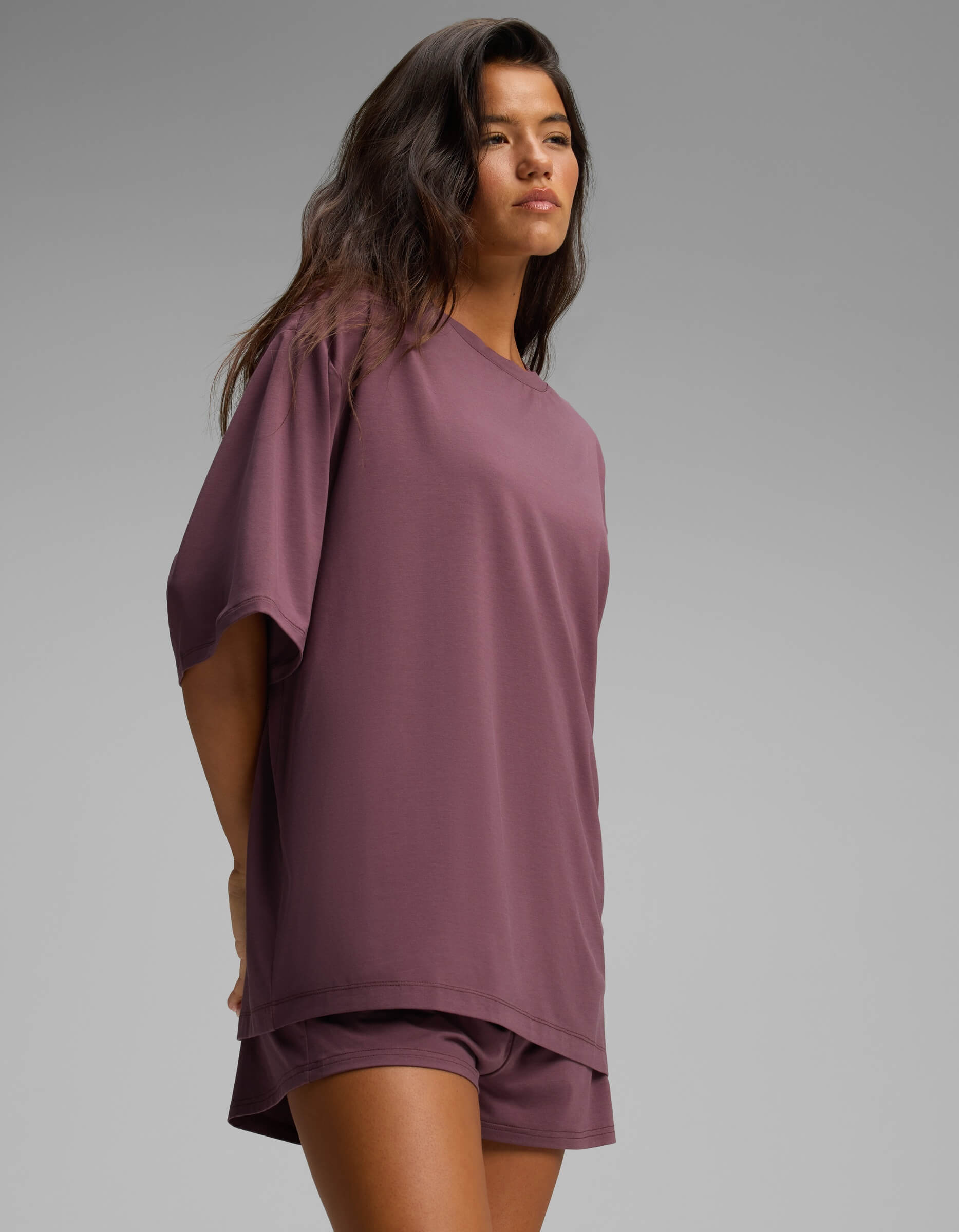 A woman with long dark hair is wearing the Cozy Earth Women’s First Layer Boyfriend Sleep Tee in purple, paired with matching shorts. She stands against a plain gray background, looking slightly to the side.