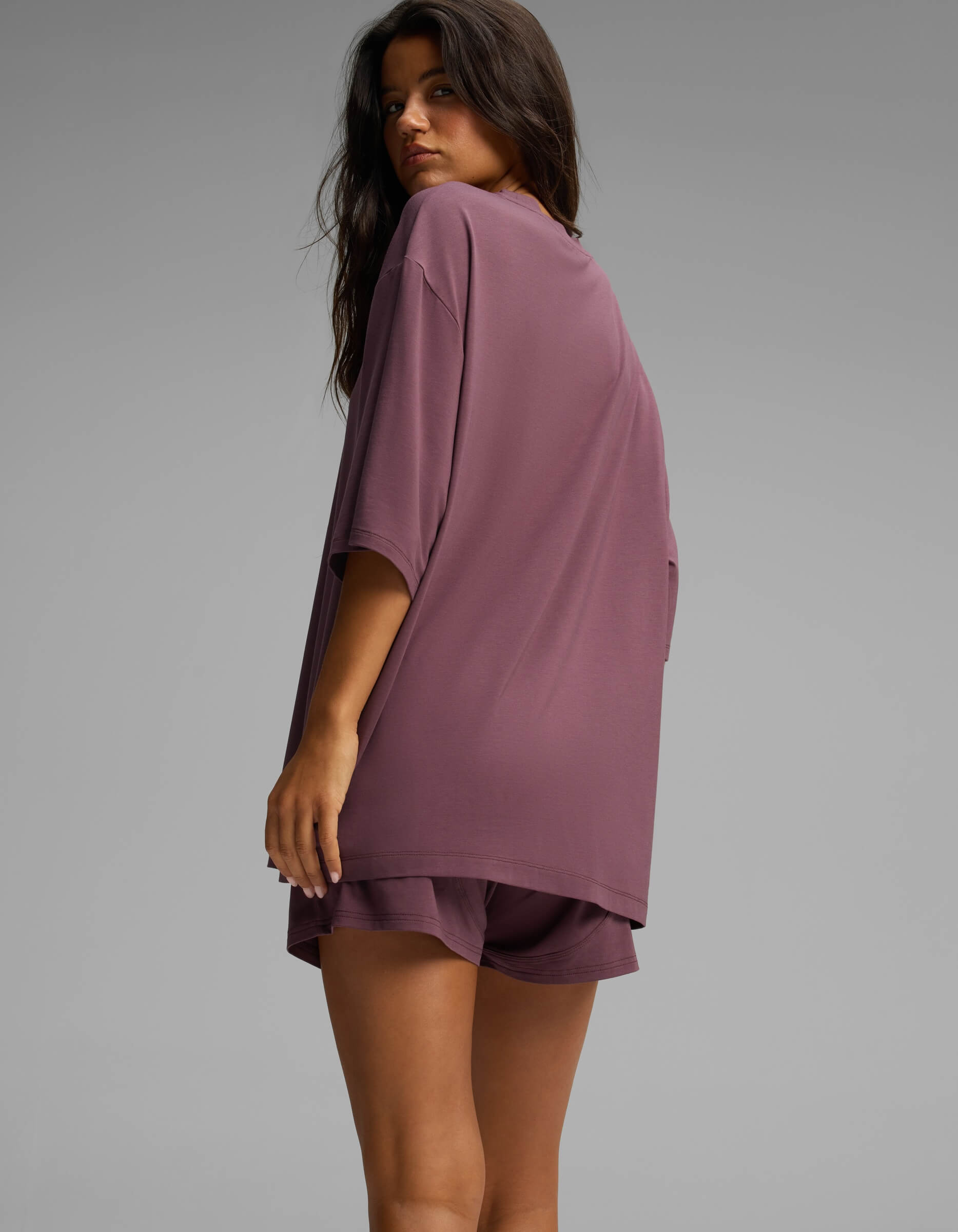 A woman with long hair stands sideways and looks over her shoulder against a plain gray background, wearing the Cozy Earth Women’s First Layer Boyfriend Sleep Tee in mauve.