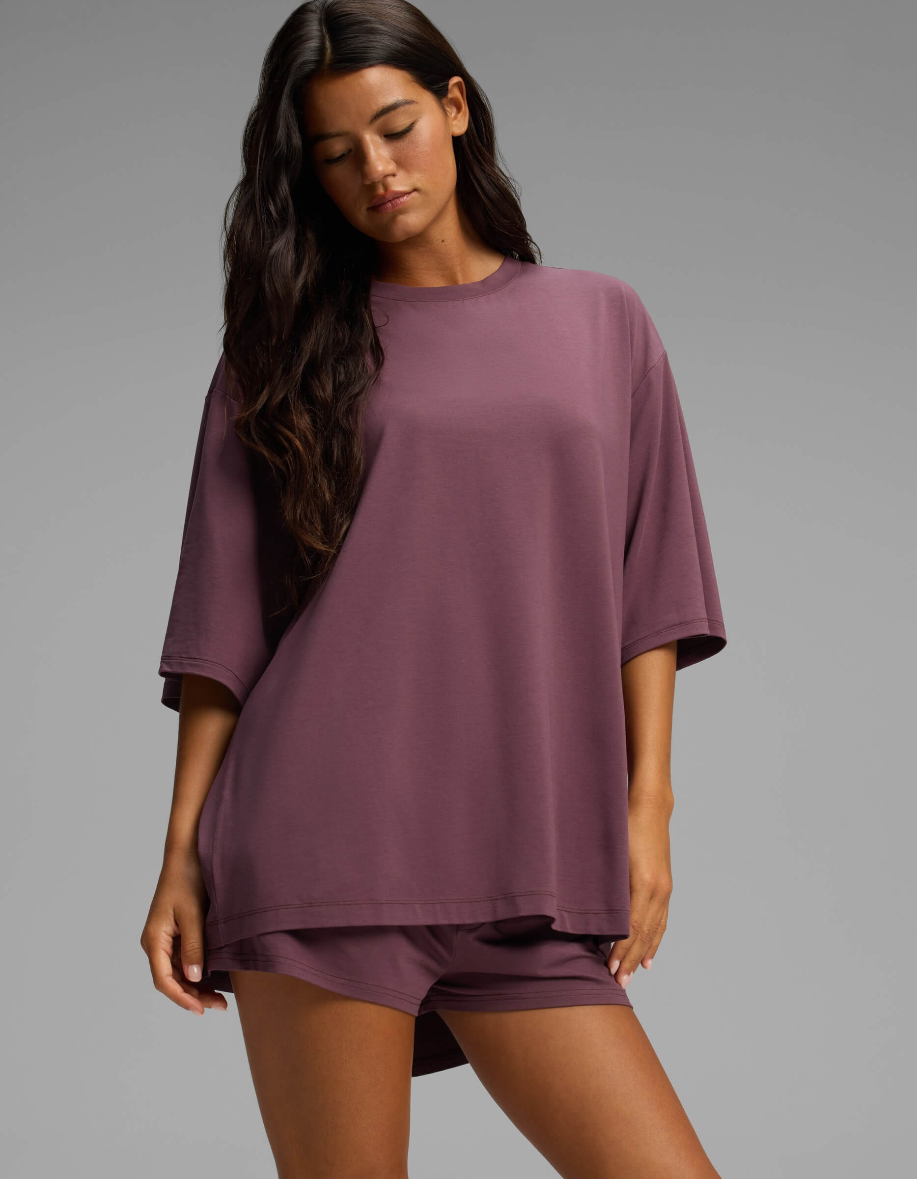 A woman with long dark hair wears the Cozy Earth Women’s First Layer Boyfriend Sleep Tee & Boxer Short Set in mauve. She looks down with a neutral expression, standing in front of a plain gray background.
