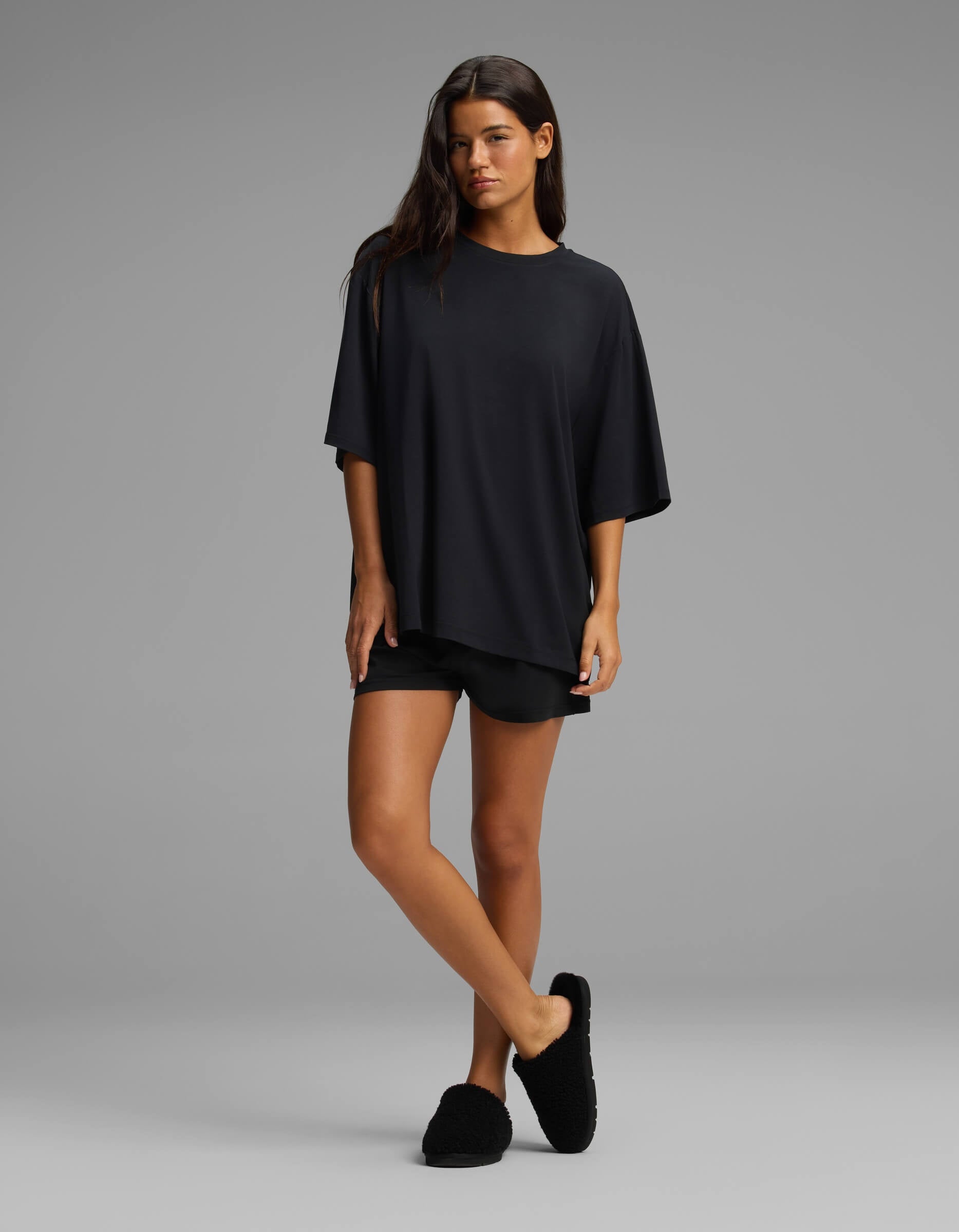 A woman with long dark hair stands against a gray background, crossing one leg in front of the other and gazing slightly to the side. She is wearing the Cozy Earth Women’s First Layer Boyfriend Sleep Tee & Boxer Short Set in black.|Color:Jet Black
