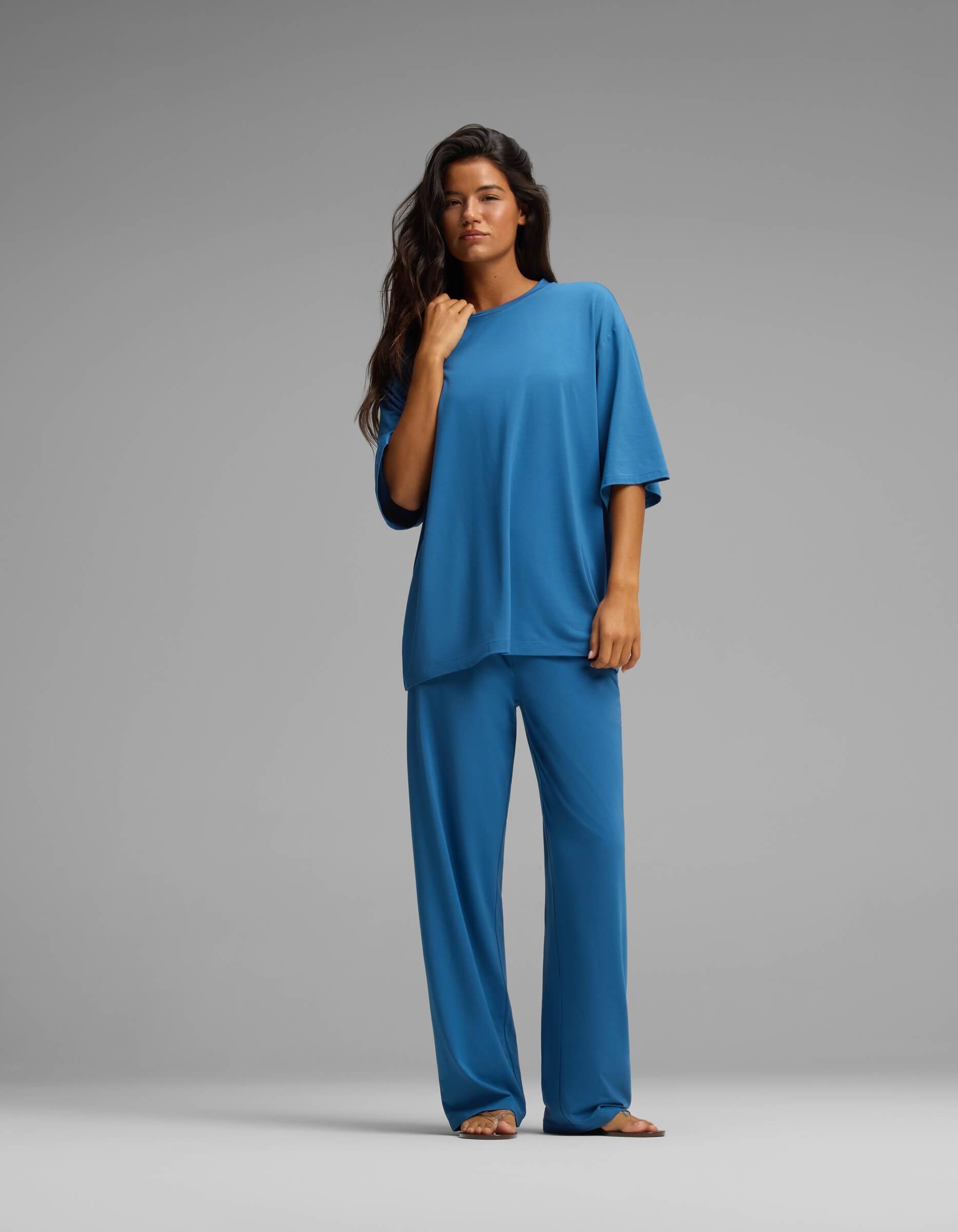 A long-haired woman stands against a grey background, wearing the Cozy Earth Women’s First Layer Boyfriend Sleep Tee & Pajama Pant Set—a loose-fitting, short-sleeved blue top paired with matching wide-leg blue pajama pants. She looks directly at the camera, one hand resting near her collarbone.