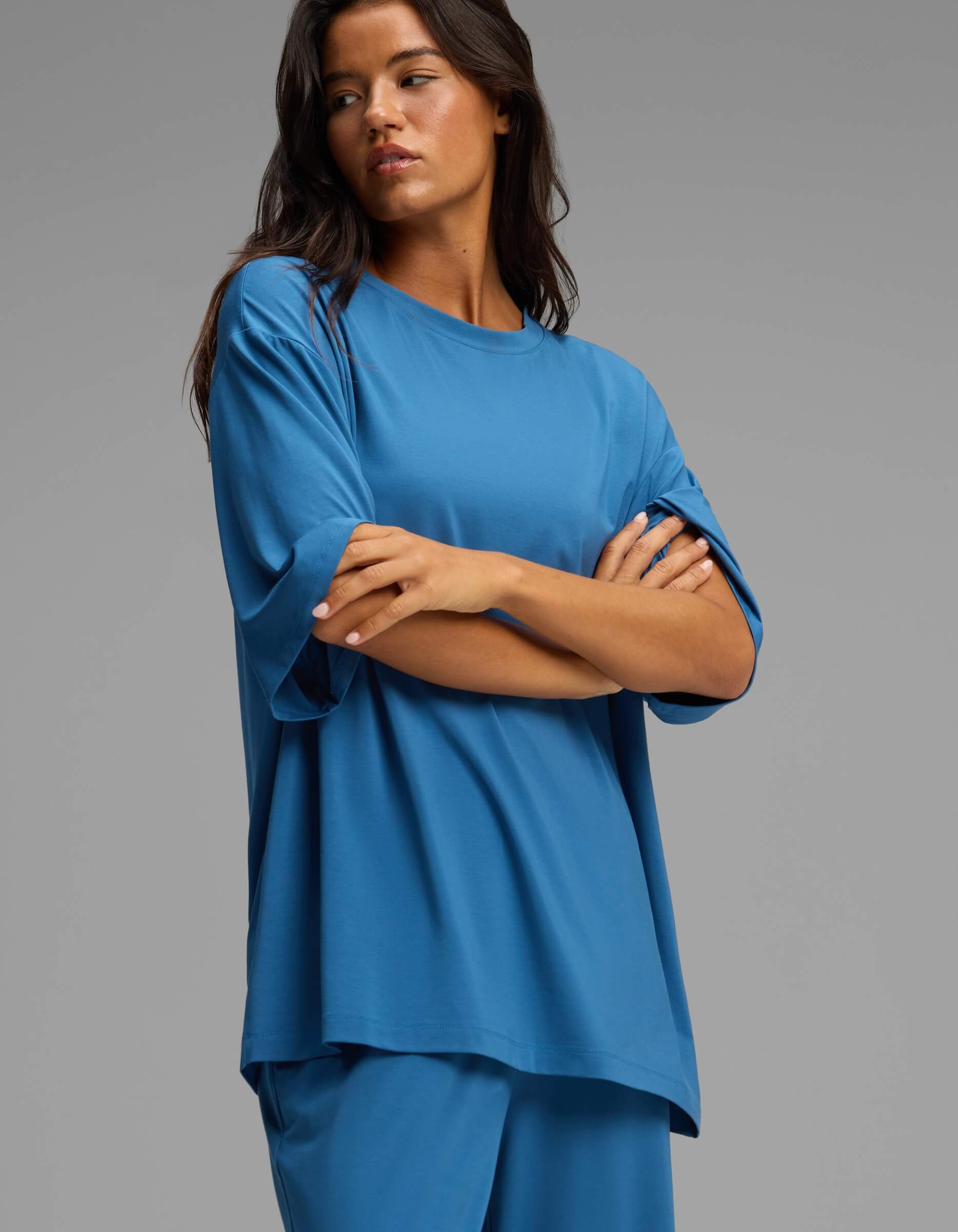 A woman with long dark hair stands with arms crossed against a plain gray background, wearing the Cozy Earth Women’s First Layer Boyfriend Sleep Tee & Pajama Pant Set in blue.