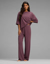 A woman stands barefoot against a gray background, wearing Cozy Earth’s Women’s First Layer Boyfriend Sleep Tee & Pajama Pant Set—a loose, mauve loungewear ensemble featuring a short-sleeve top and relaxed pants.|Color:Fig