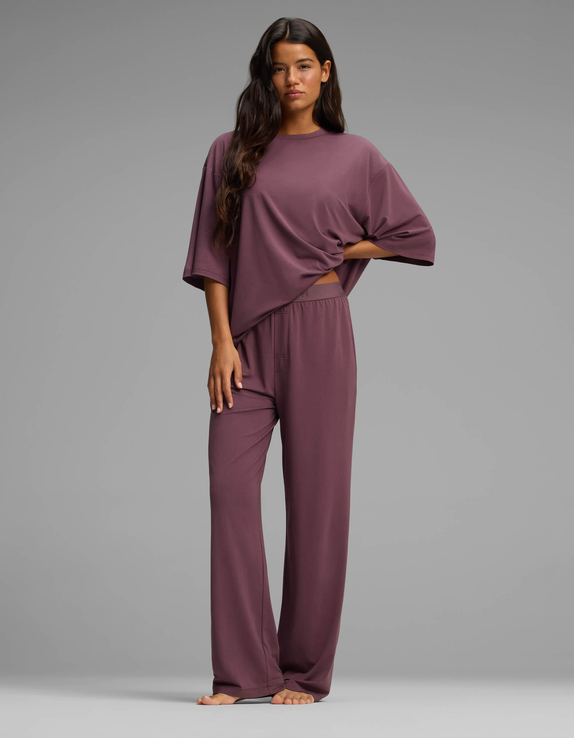 A woman stands barefoot against a gray background, wearing Cozy Earth’s Women’s First Layer Boyfriend Sleep Tee & Pajama Pant Set—a loose, mauve loungewear ensemble featuring a short-sleeve top and relaxed pants.