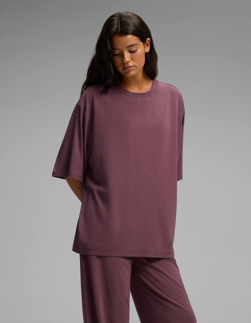 A woman with long dark hair stands against a plain gray background, her hands behind her back, wearing the Cozy Earth Women’s First Layer Boyfriend Sleep Tee & Pajama Pant Set in a loose mauve fit.