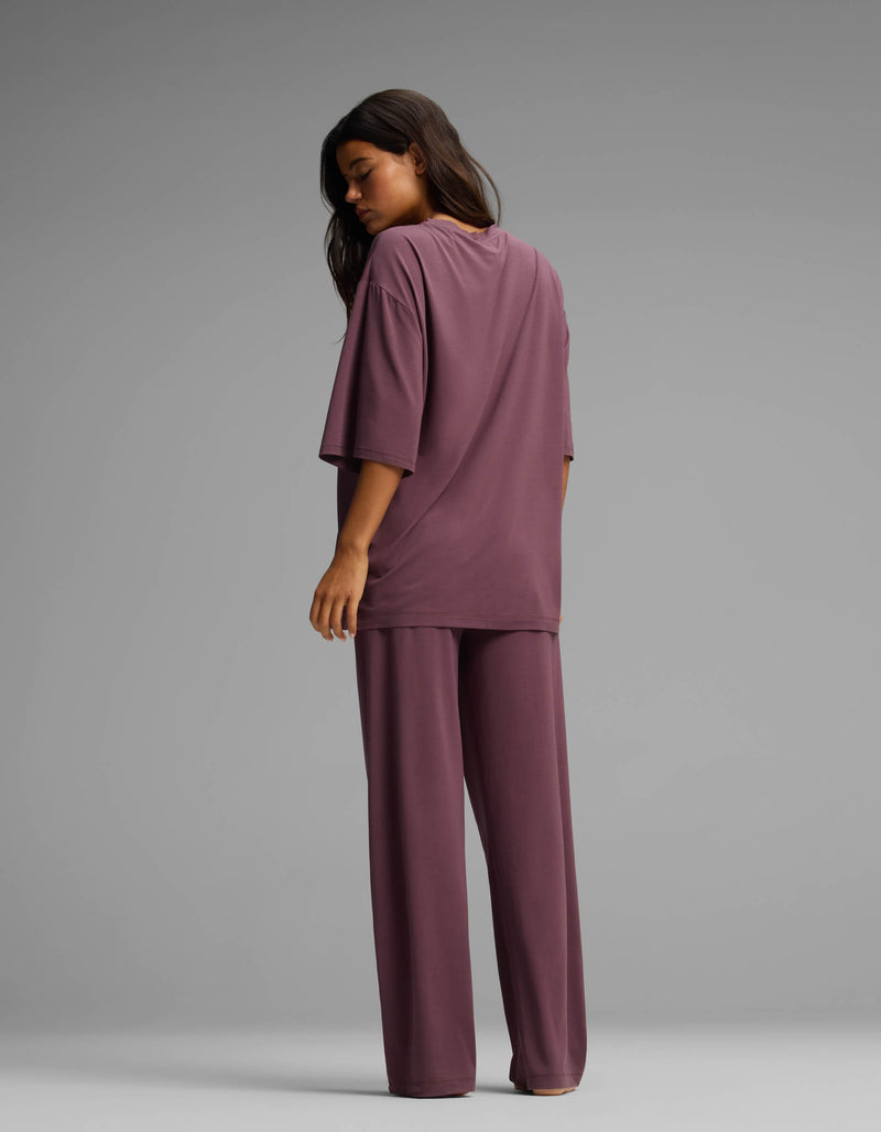 A woman stands facing away, wearing the Cozy Earth Women’s First Layer Boyfriend Sleep Tee & Pajama Pant Set—a loose-fitting, mauve-colored short-sleeve top and matching wide-legged pants—against a plain gray background.