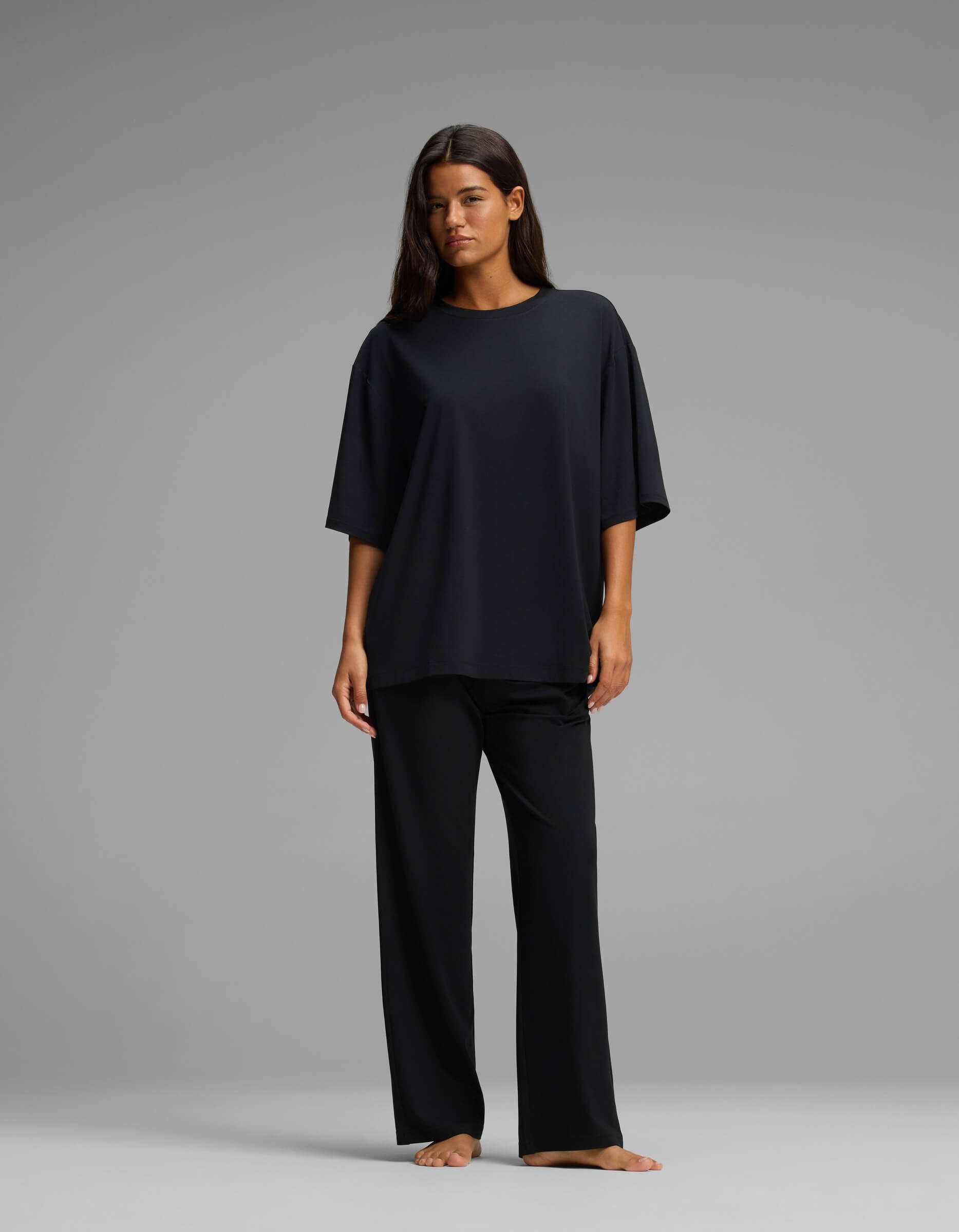 A woman stands barefoot against a gray background, wearing the Cozy Earth Women’s First Layer Boyfriend Sleep Tee & Pajama Pant Set in black, with her hair down and her arms relaxed by her sides.|Color:Jet Black