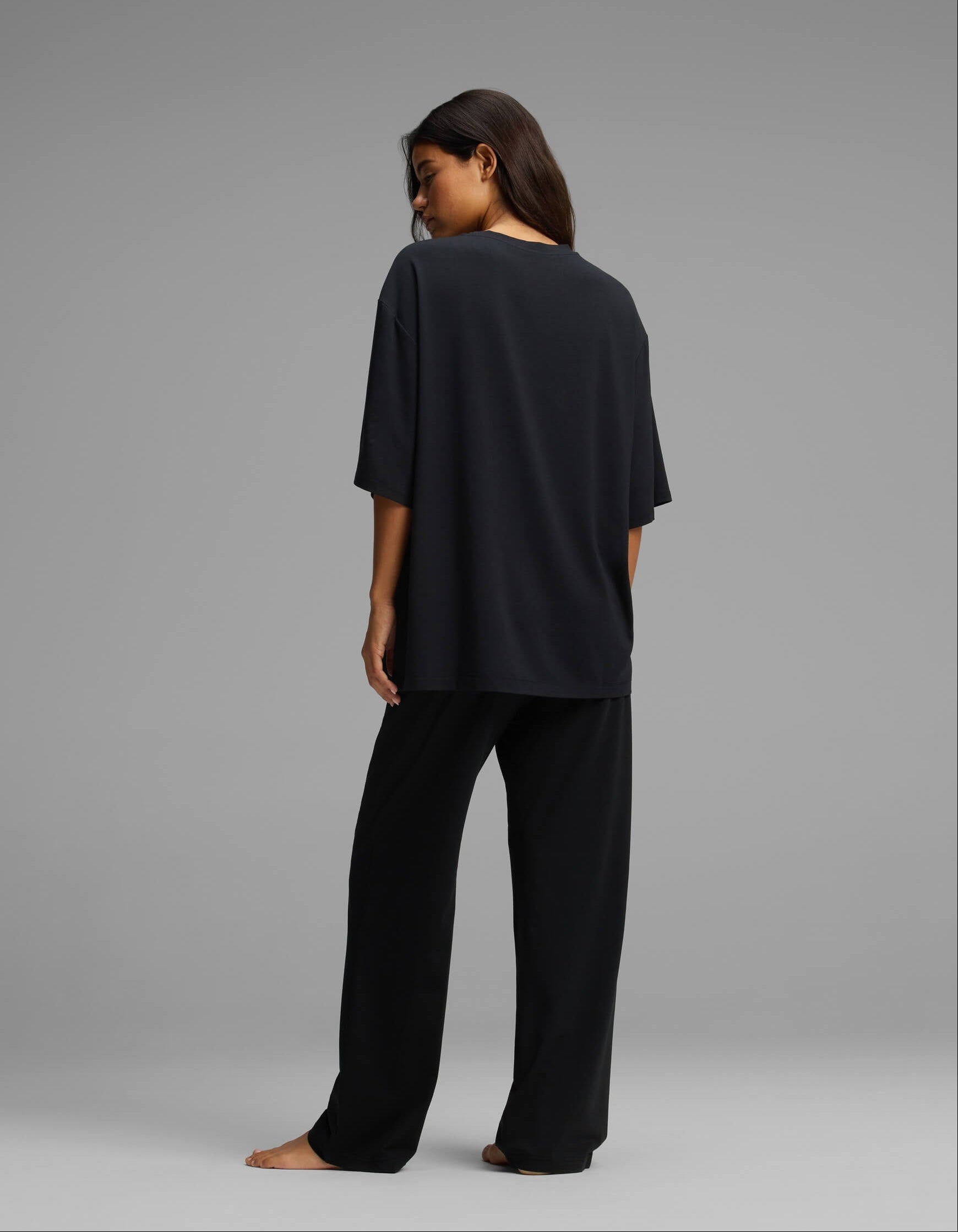 A woman stands barefoot, facing away, wearing the Cozy Earth Women’s First Layer Boyfriend Sleep Tee & Pajama Pant Set in black against a plain gray background.