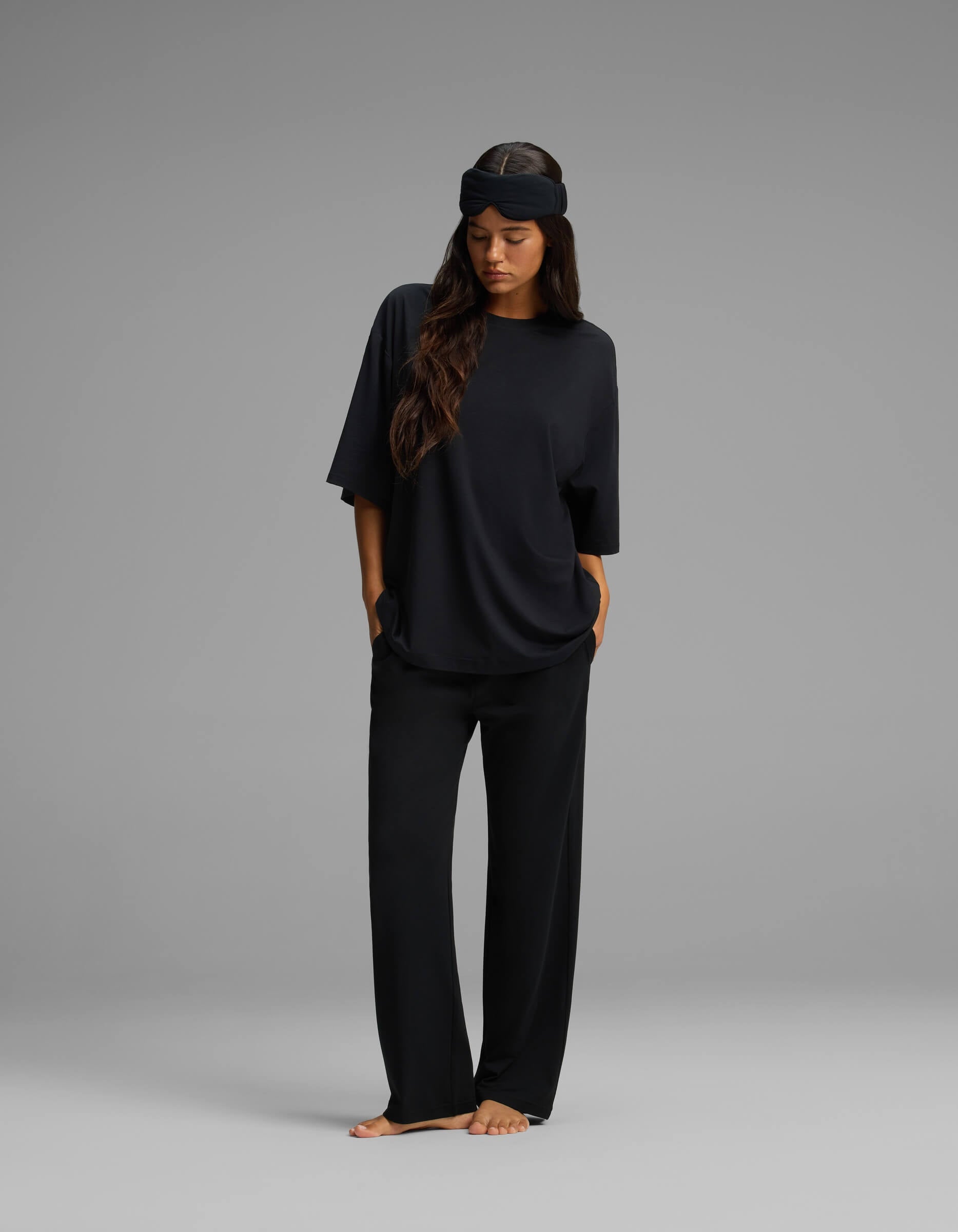 A woman stands barefoot against a gray background, wearing the Cozy Earth Women’s First Layer Boyfriend Sleep Tee & Pajama Pant Set in black. The oversized, relaxed-fit T-shirt and loose pajama pants exude comfort, while a black sleep mask is pushed up on her forehead. Her hands rest in her pockets and her long hair drapes over one shoulder.