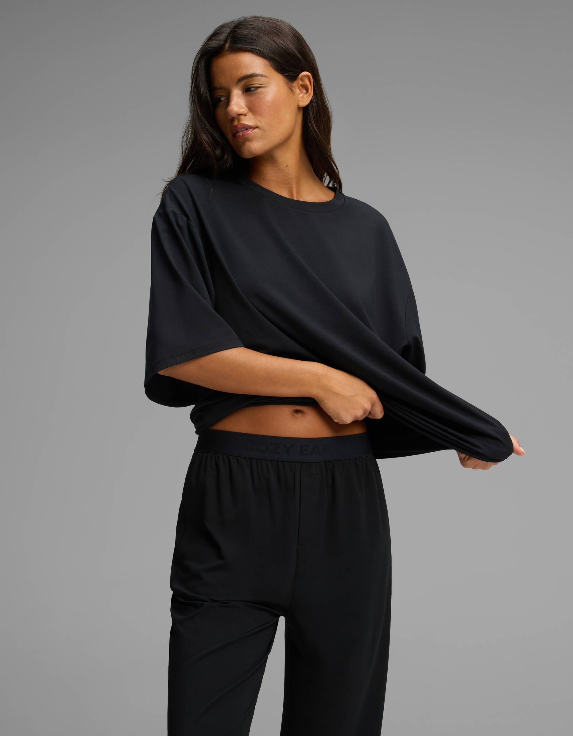 A woman models the Cozy Earth Women’s First Layer Boyfriend Sleep Tee & Pajama Pant Set in black, standing against a plain gray background and gently lifting her shirt to reveal the waistband of the matching pants as she looks off to the side.