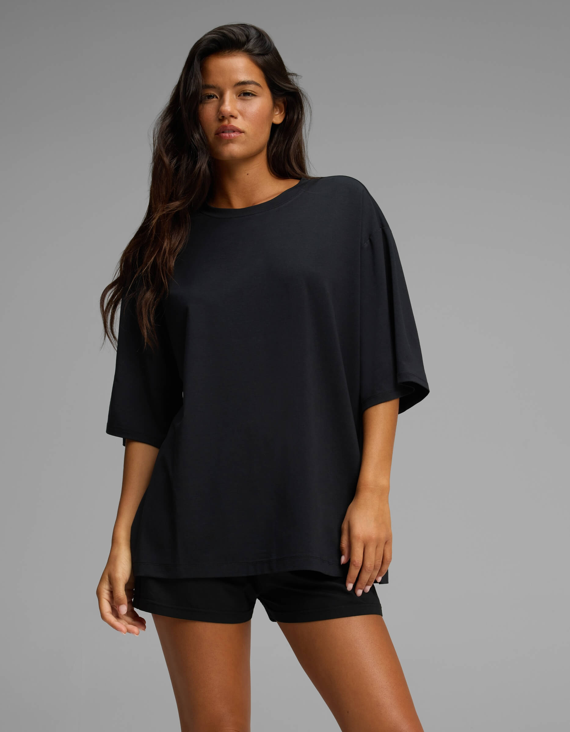 A woman with long dark hair models the Cozy Earth Women’s First Layer Boyfriend Sleep Tee in black, paired with matching shorts, as she stands confidently against a plain gray background.