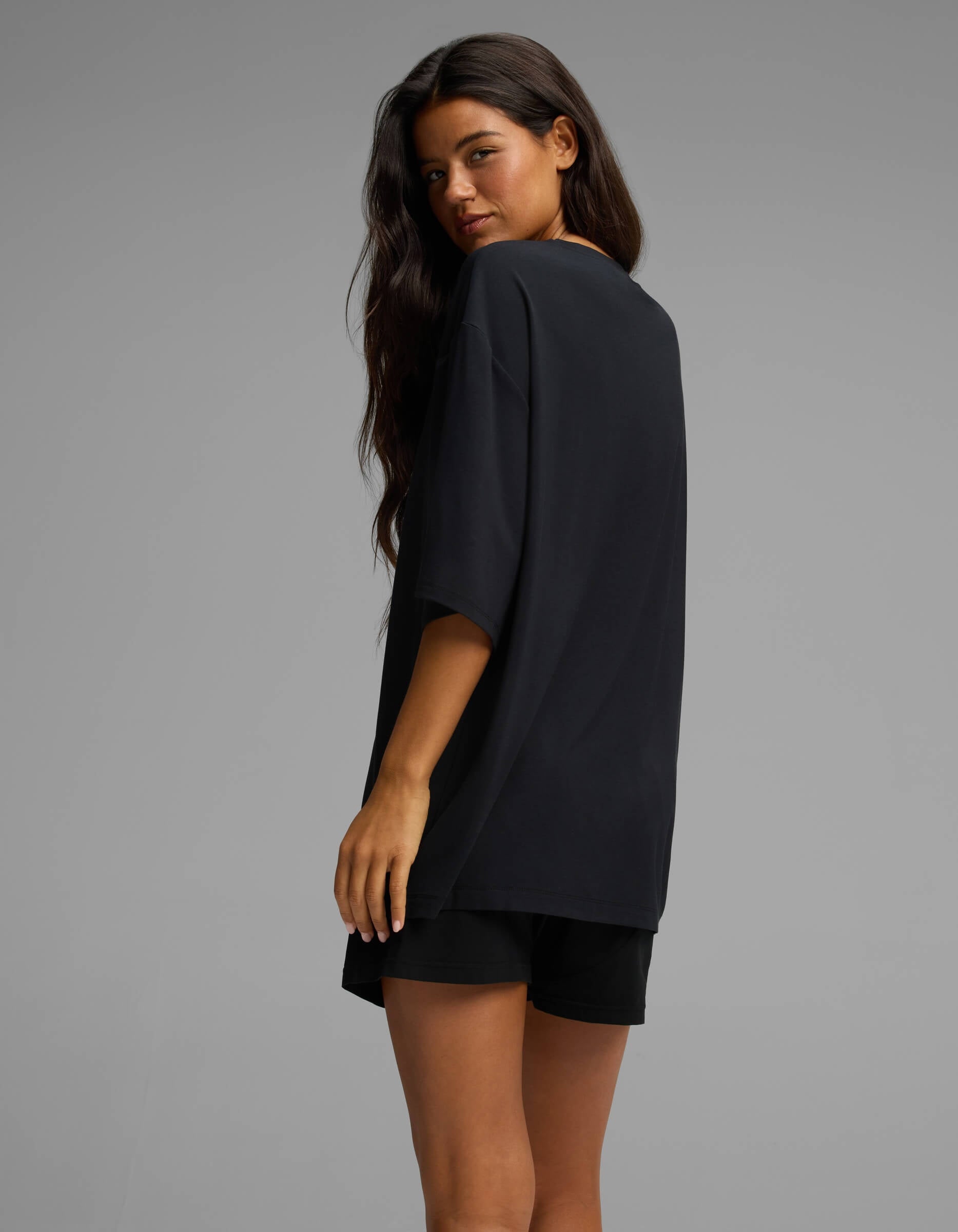 A woman with long dark hair wearing the Cozy Earth Women’s First Layer Boyfriend Sleep Tee in black stands turned slightly to the side, looking over her shoulder against a plain gray background.