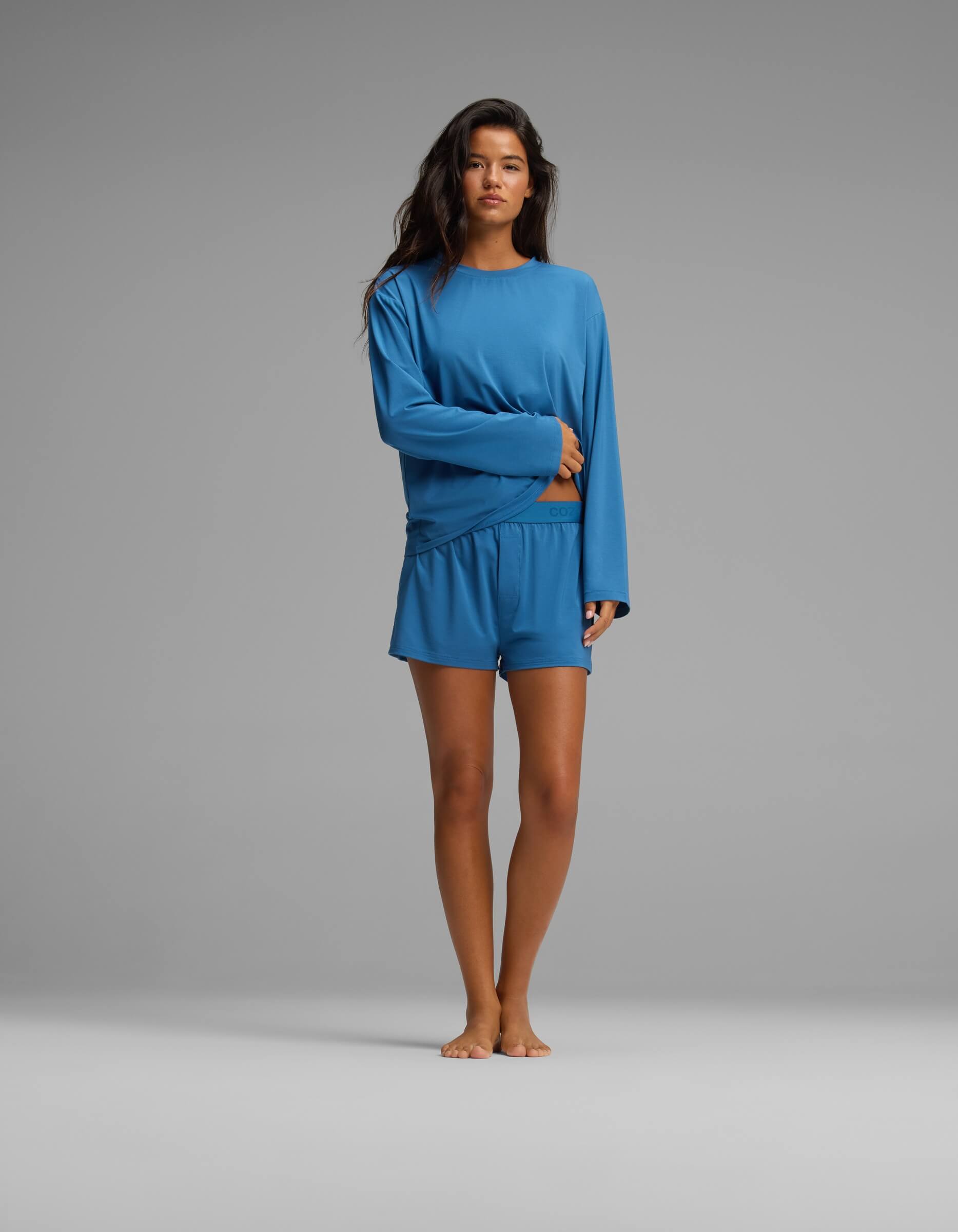 A woman stands barefoot on a plain background, wearing the Cozy Earth Women’s First Layer Long Sleeve Sleep Tee in blue with matching blue shorts.
