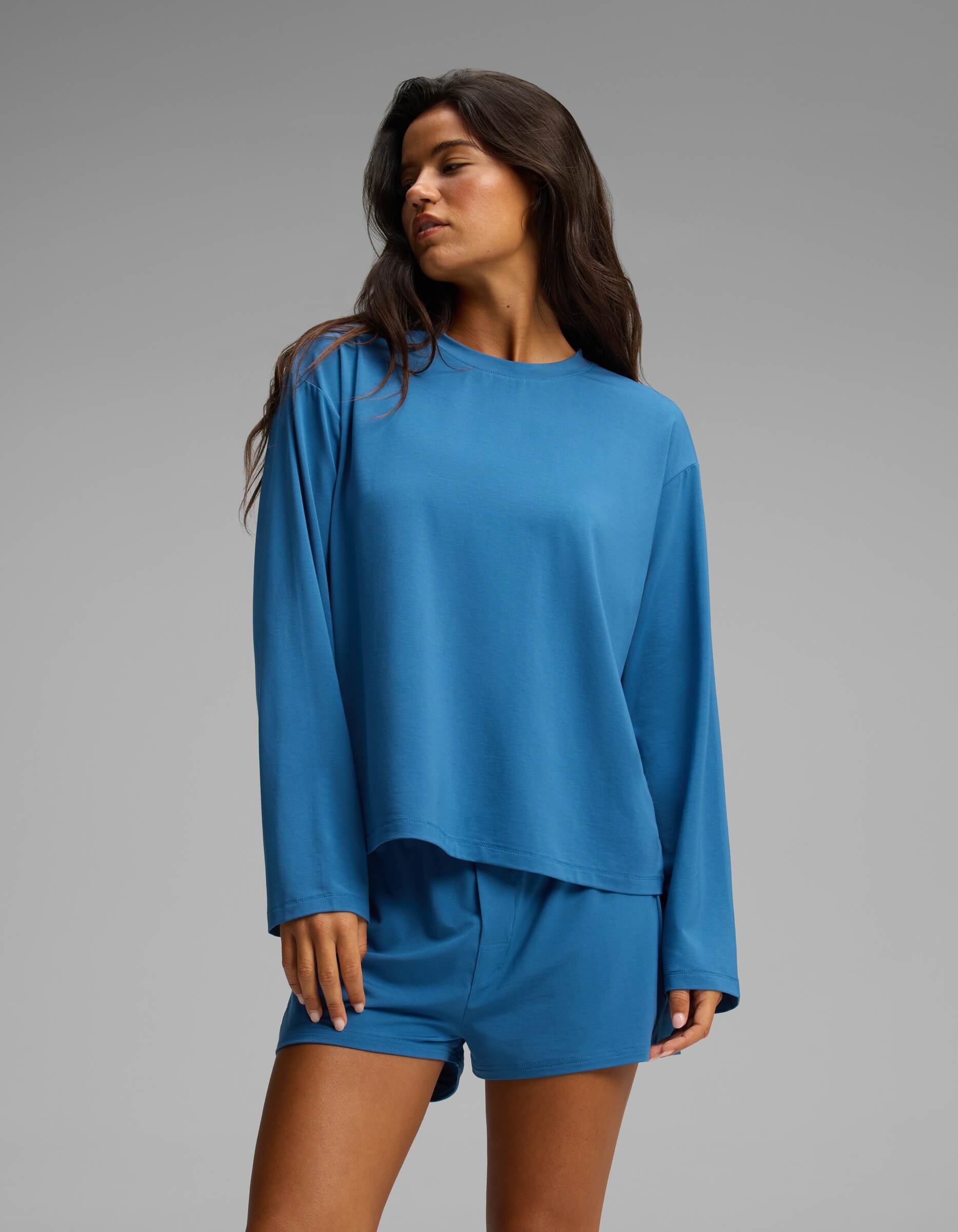 A woman wearing the Cozy Earth Women’s First Layer Long Sleeve Sleep Tee in blue and coordinating shorts stands against a plain gray background, looking to the side with her hand relaxed by her leg.|Color:Anchor