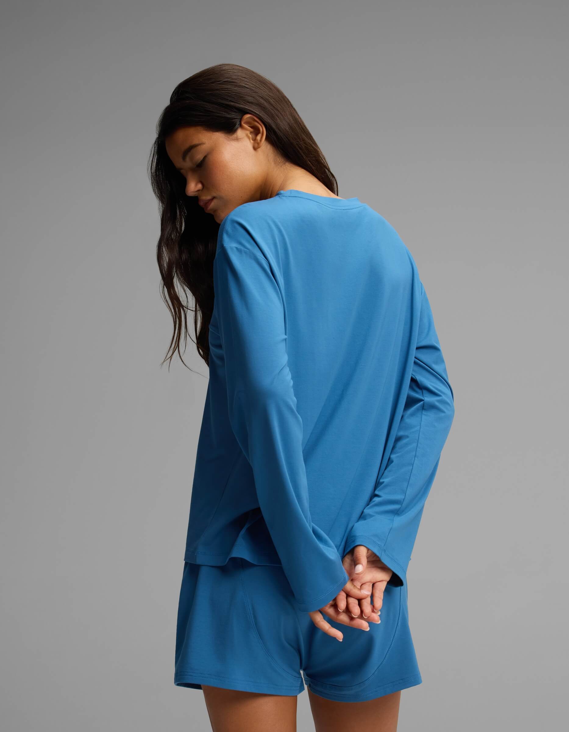 A woman with long dark hair wears the Cozy Earth Women’s First Layer Long Sleeve Sleep Tee in blue, paired with matching shorts. She stands against a plain gray background with her hands clasped behind her back, looking downward.