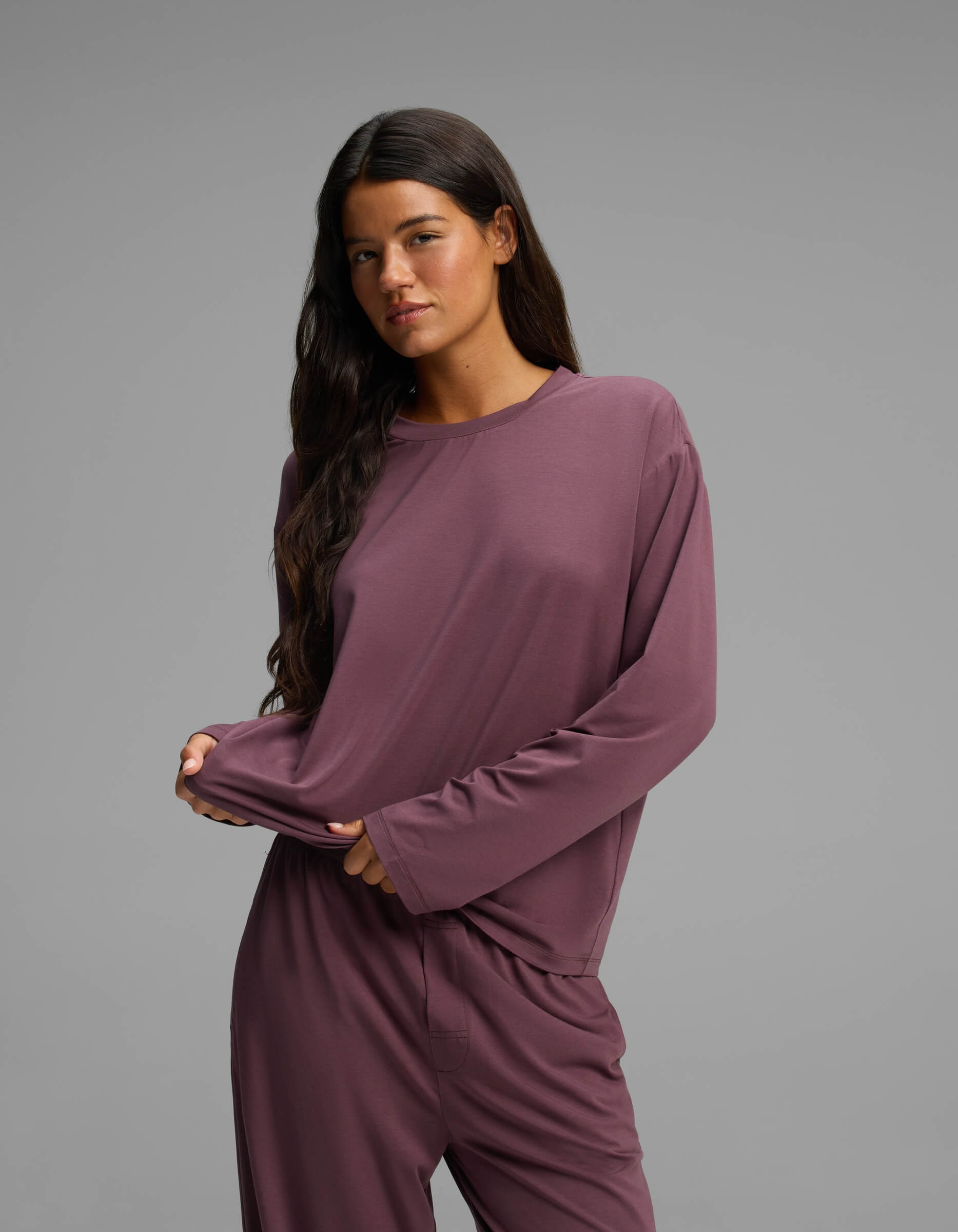 A woman with long dark hair stands against a plain gray background, gently pulling at her shirt with one hand. She is wearing the Cozy Earth Women’s First Layer Long Sleeve Sleep Tee and matching pants in mauve.