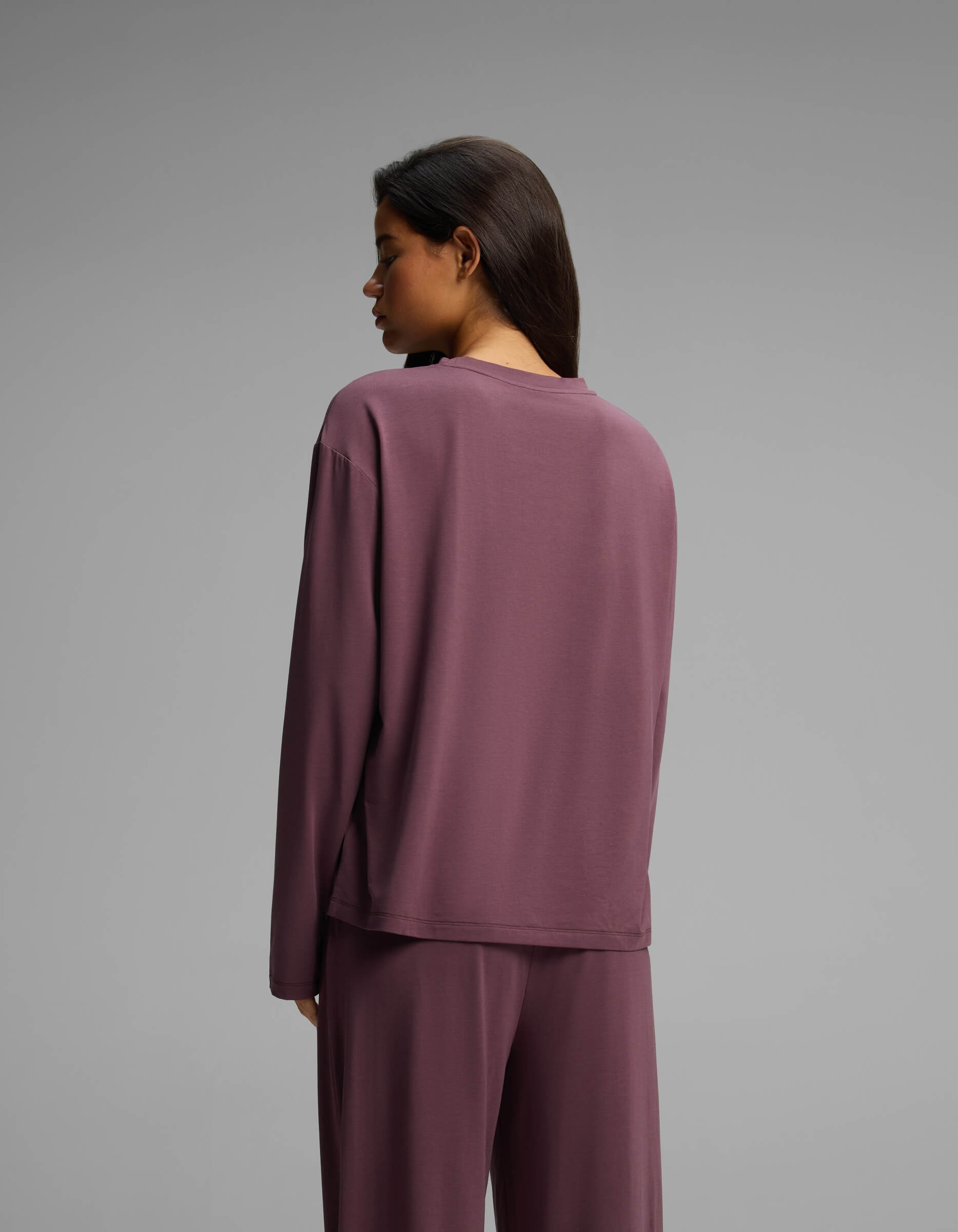 A woman with long dark hair, wearing the Cozy Earth Women’s First Layer Long Sleeve Sleep Tee and matching pants in mauve, stands facing away from the camera against a plain gray background.