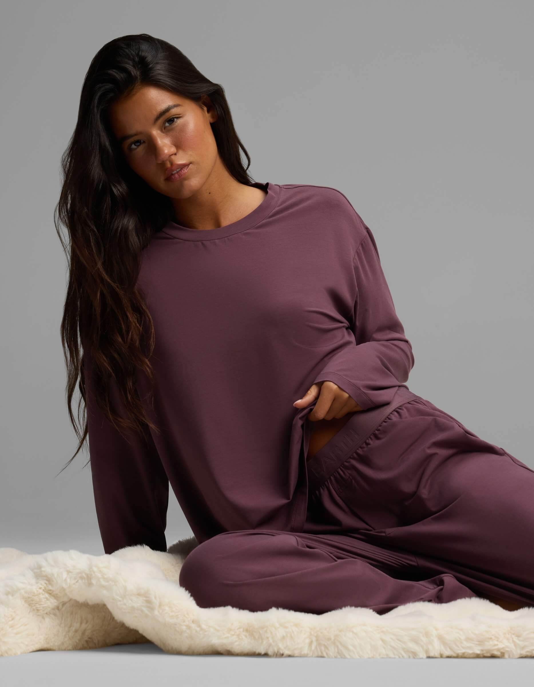 A woman with long dark hair sits on a cream-colored rug, wearing the Cozy Earth Women’s First Layer Long Sleeve Sleep Tee in mauve with matching pants, looking at the camera with a neutral expression.