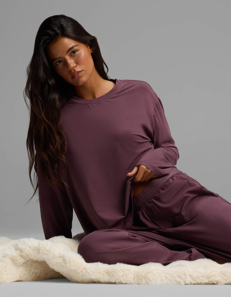 A woman with long dark hair sits on a cream-colored rug, wearing the Cozy Earth Women’s First Layer Long Sleeve Sleep Tee in mauve with matching pants, looking at the camera with a neutral expression.