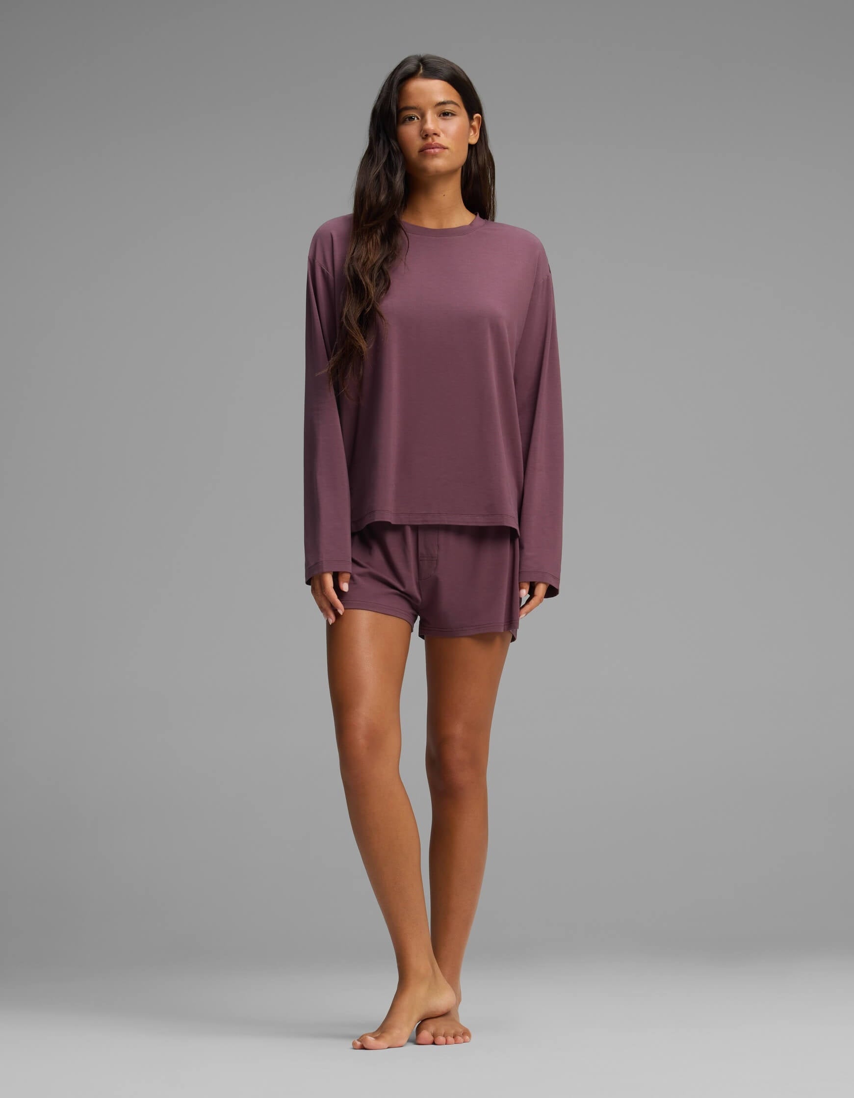 A woman stands barefoot against a plain gray background, wearing the Cozy Earth Women’s First Layer Long Sleeve Sleep Tee & Boxer Short Set in mauve, her long dark hair draped over her shoulder.|Color:Fig