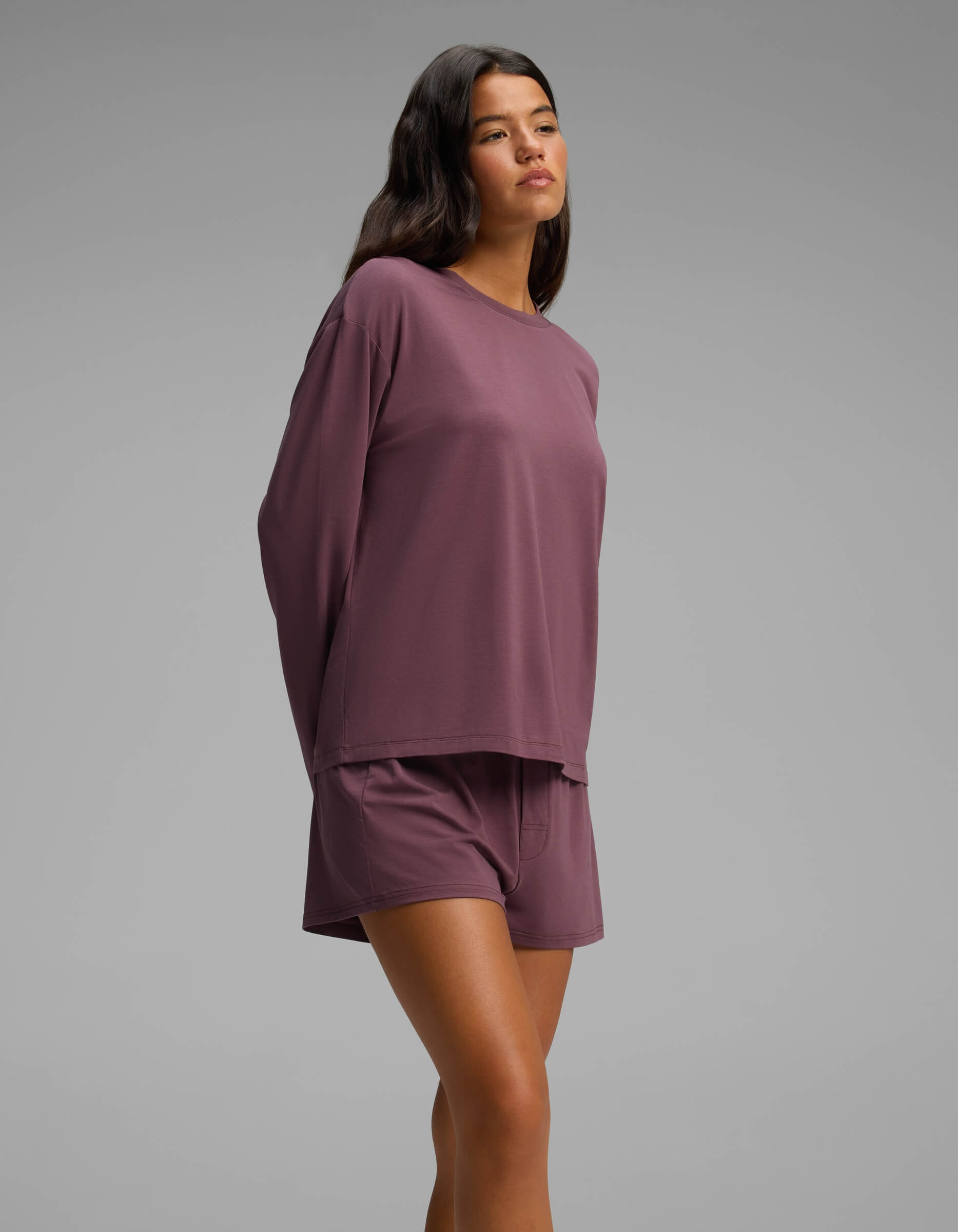 Dressed in the Cozy Earth Women’s First Layer Long Sleeve Sleep Tee & Boxer Short Set, a woman stands against a plain gray background with her hands behind her back, gazing slightly upward.