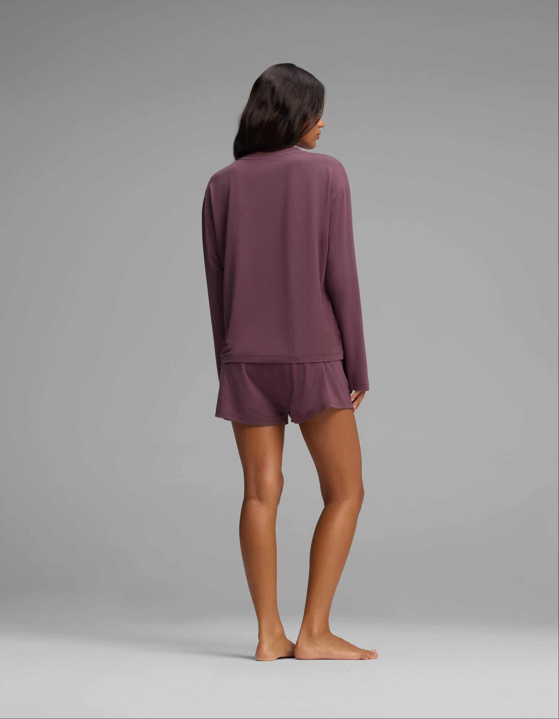A woman stands barefoot facing away, wearing the Cozy Earth Women’s First Layer Long Sleeve Sleep Tee & Boxer Short Set in mauve, against a plain gray background.