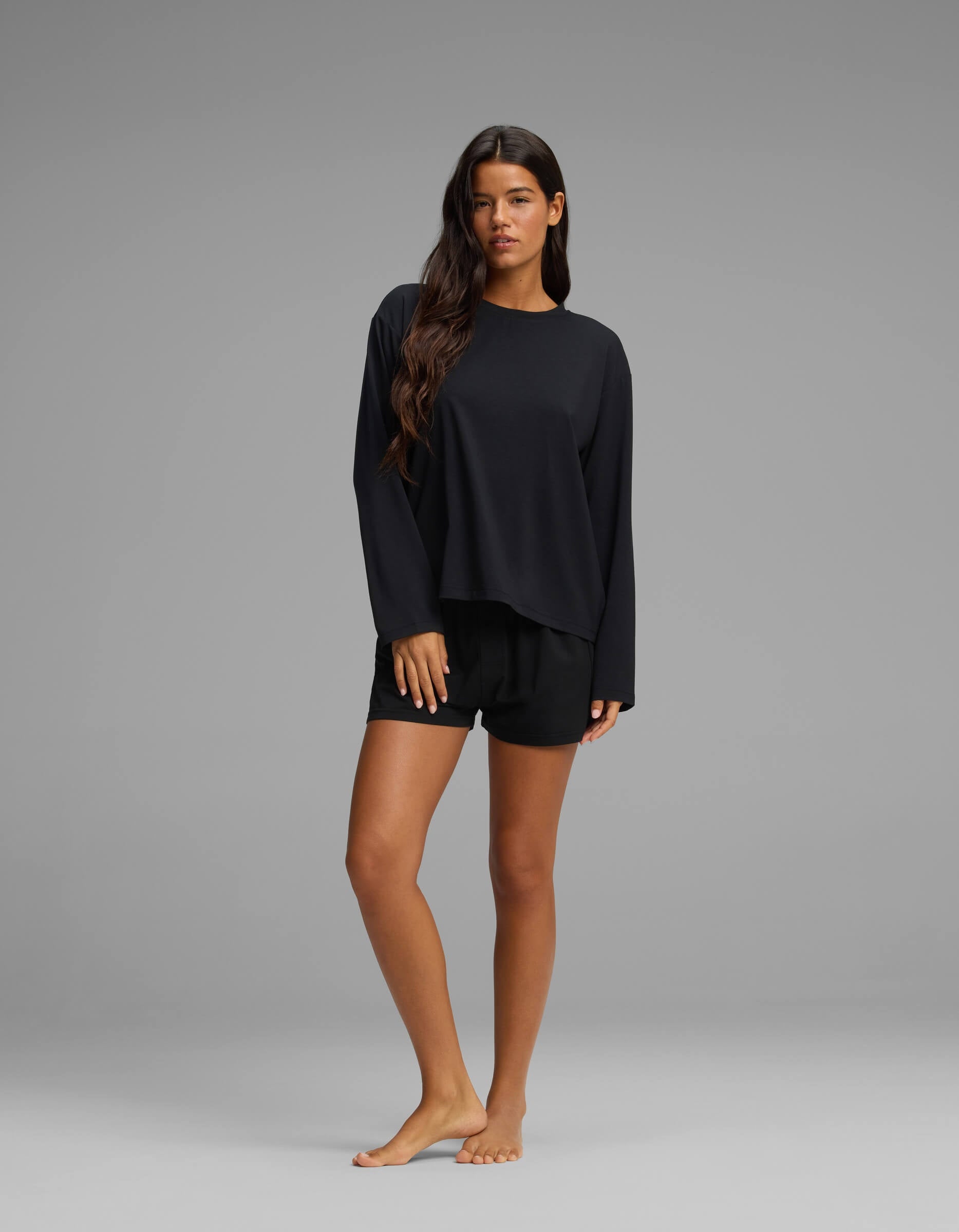 A woman with long brown hair stands barefoot on a grey background, modeling the Cozy Earth Women’s First Layer Long Sleeve Sleep Tee & Boxer Short Set in black, looking at the camera with a neutral expression.|Color:Jet Black