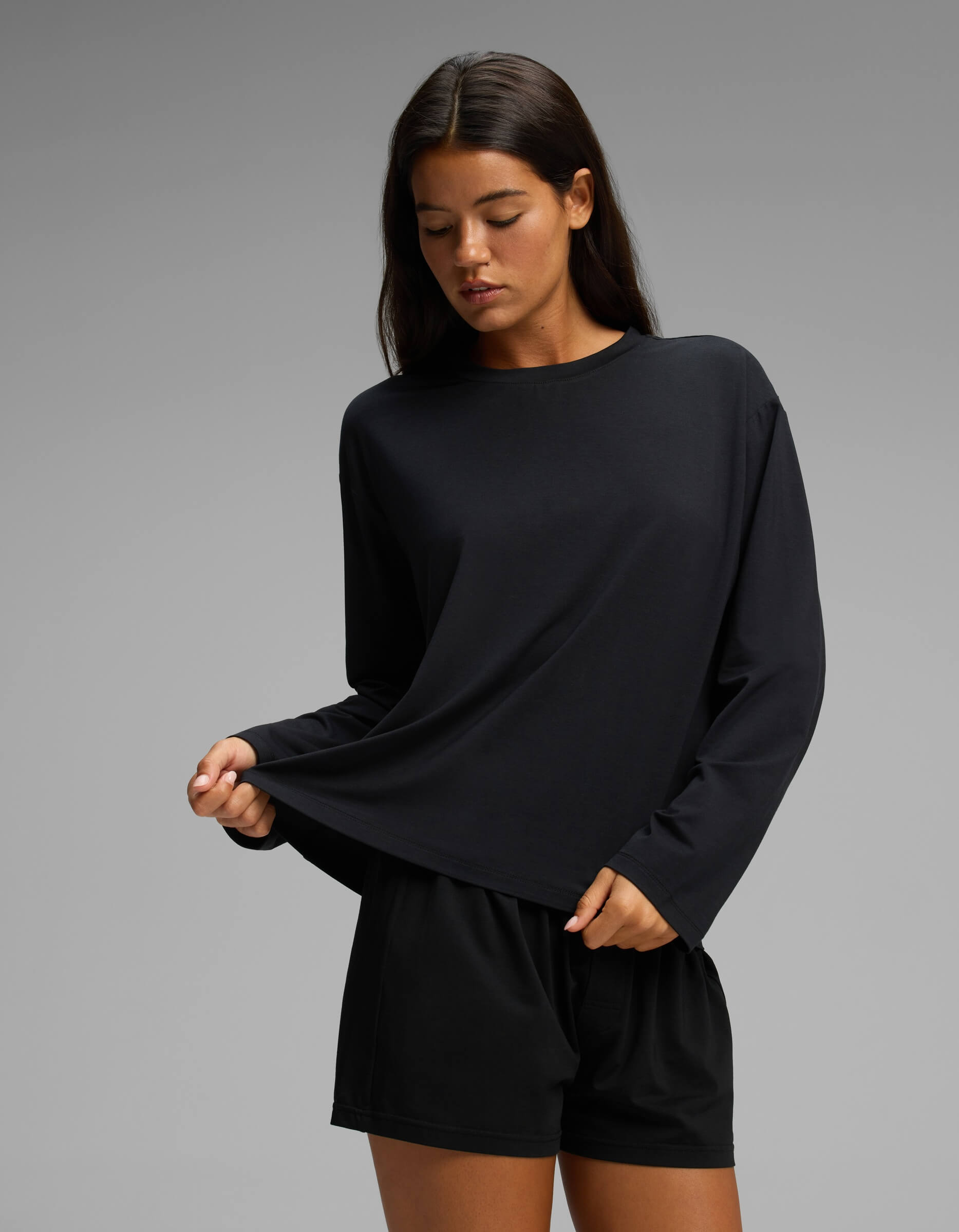 A woman stands against a plain gray background, gently pulling at the hem of her black Cozy Earth Women’s First Layer Long Sleeve Sleep Tee & Boxer Short Set while looking down.
