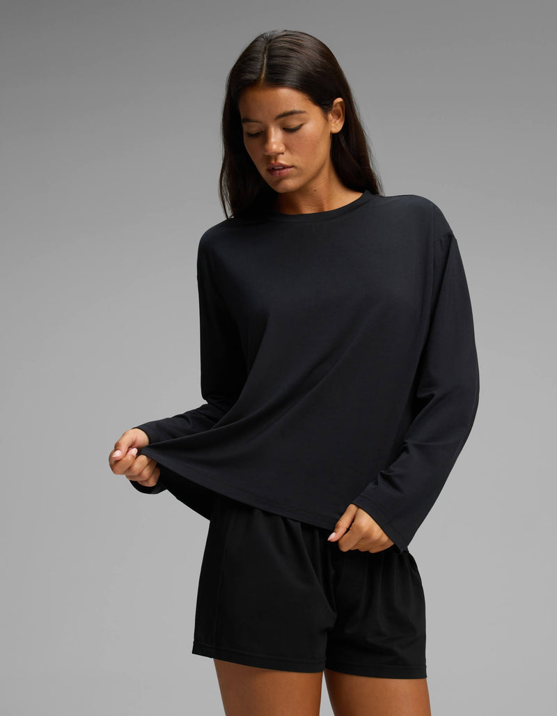 A woman stands against a plain gray background, gently pulling at the hem of her black Cozy Earth Women’s First Layer Long Sleeve Sleep Tee & Boxer Short Set while looking down.