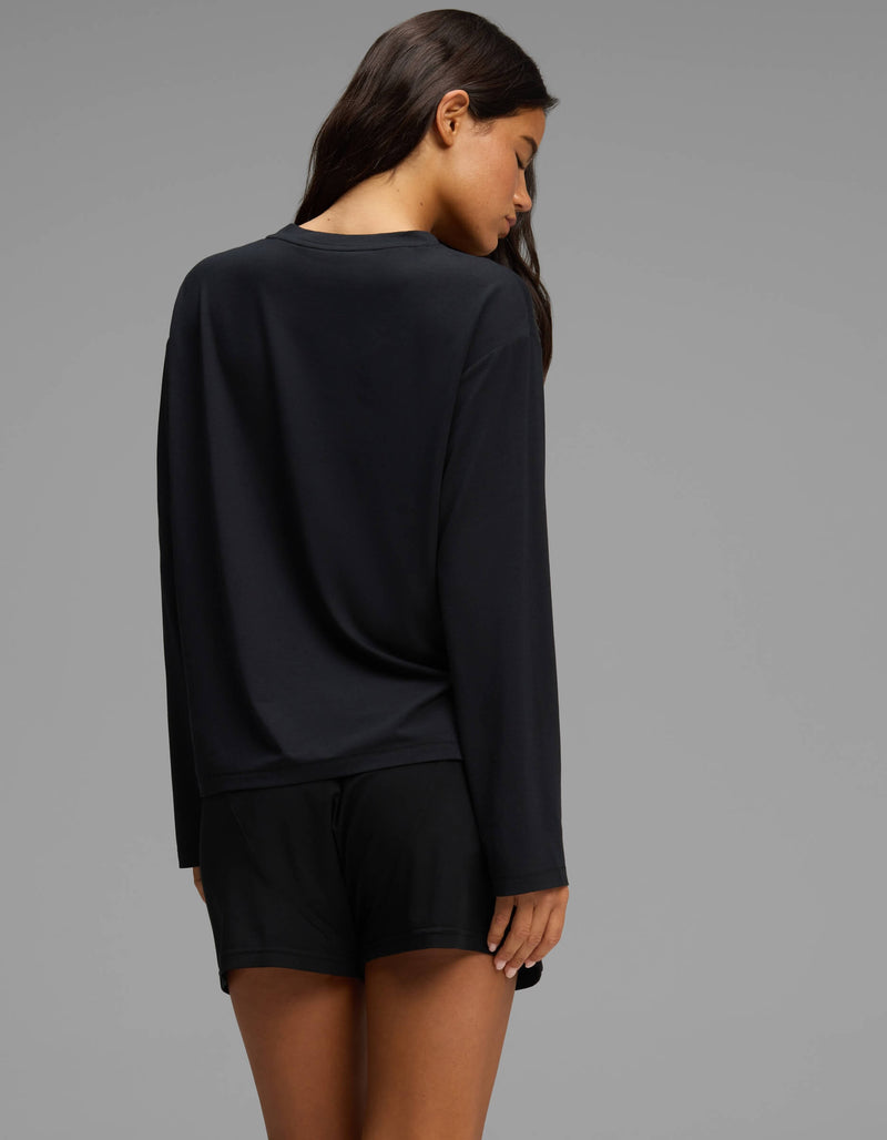 A woman with long brown hair is seen from behind, looking down, wearing the Cozy Earth Women’s First Layer Long Sleeve Sleep Tee & Boxer Short Set in black against a plain gray background.