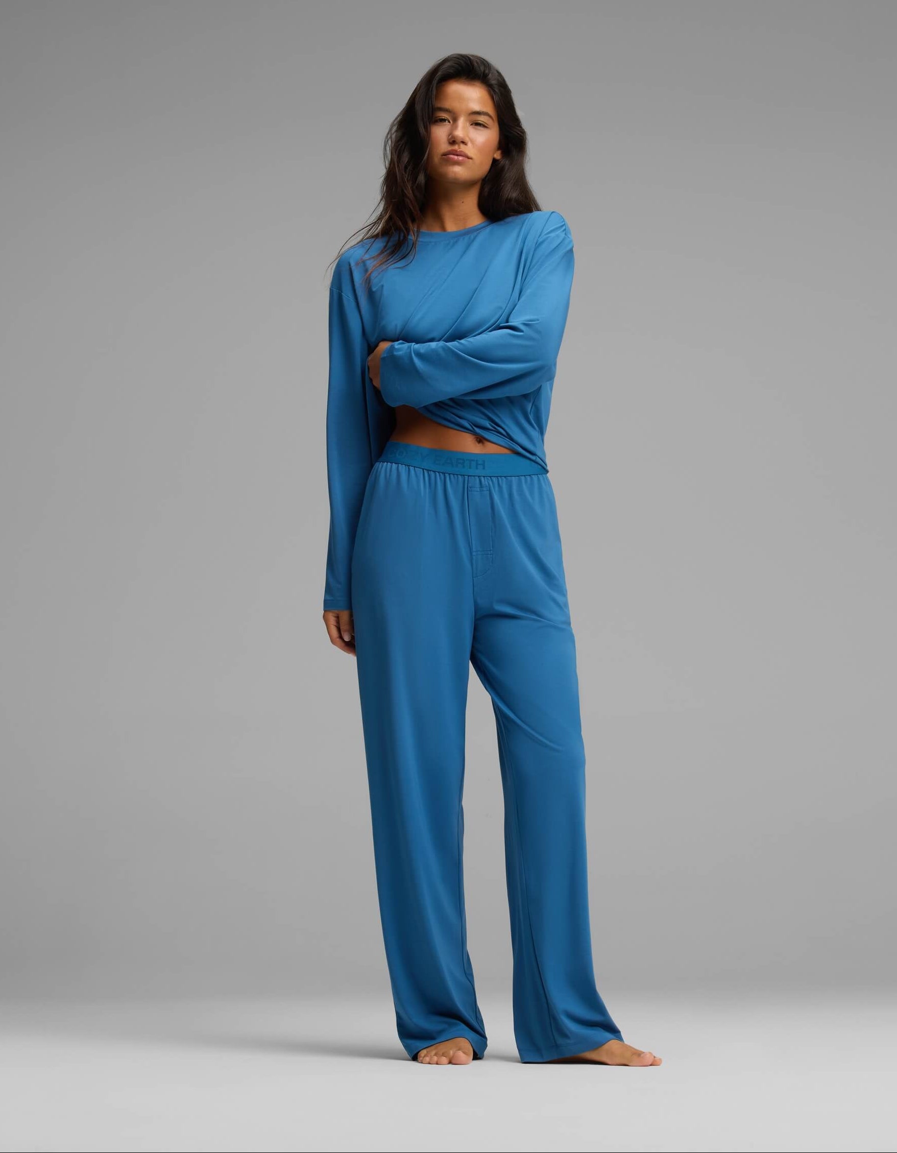 A person stands barefoot against a plain gray background, wearing the Cozy Earth Women’s First Layer Long Sleeve Sleep Tee & Pajama Pant Set in blue. They have long dark hair and a neutral expression, with one arm crossed over their body.|Color:Anchor