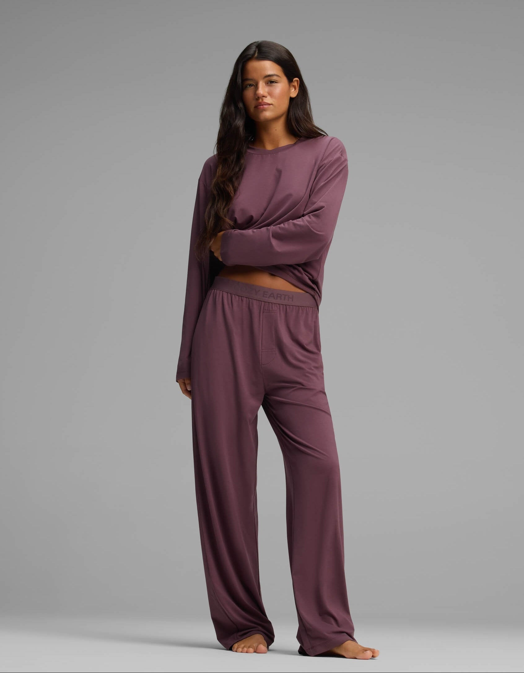 A woman with long, dark hair stands barefoot against a gray background, arms crossed and wearing the Cozy Earth Women’s First Layer Long Sleeve Sleep Tee & Pajama Pant Set in mauve. The loose-fitting pajama set features matching long-sleeved top and pants, and she has a neutral expression.|Color:Fig