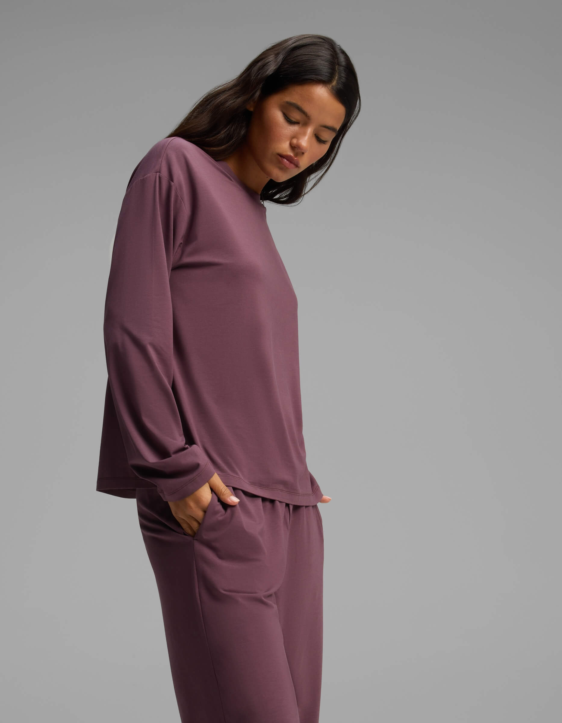 A woman with dark hair stands against a gray backdrop, wearing the Cozy Earth Women’s First Layer Long Sleeve Sleep Tee & Pajama Pant Set in a rich plum shade. She has her hands tucked into the pockets of her comfortable pajama pants and is looking downward.