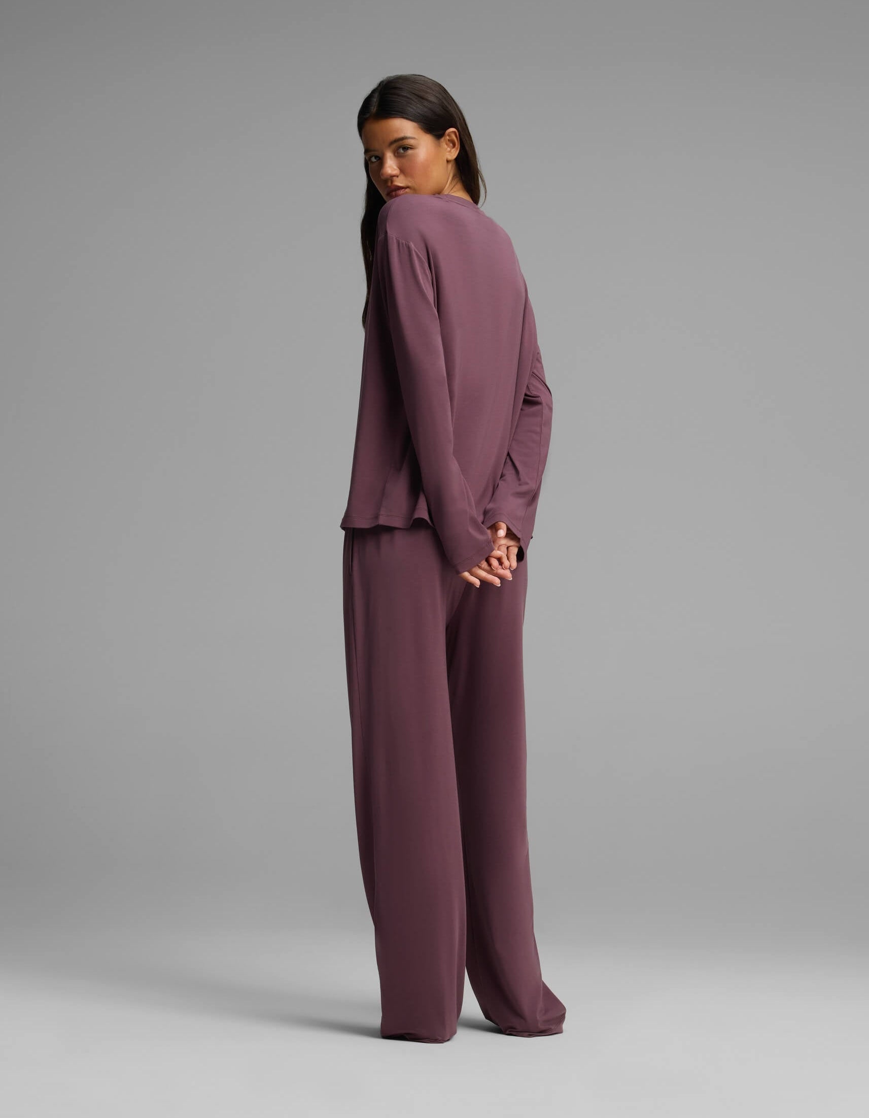 A woman stands with her back partially turned against a plain gray background, her hands clasped behind her. She is wearing the Cozy Earth Women’s First Layer Long Sleeve Sleep Tee & Pajama Pant Set in mauve, featuring a loose fit and wide-leg silhouette.